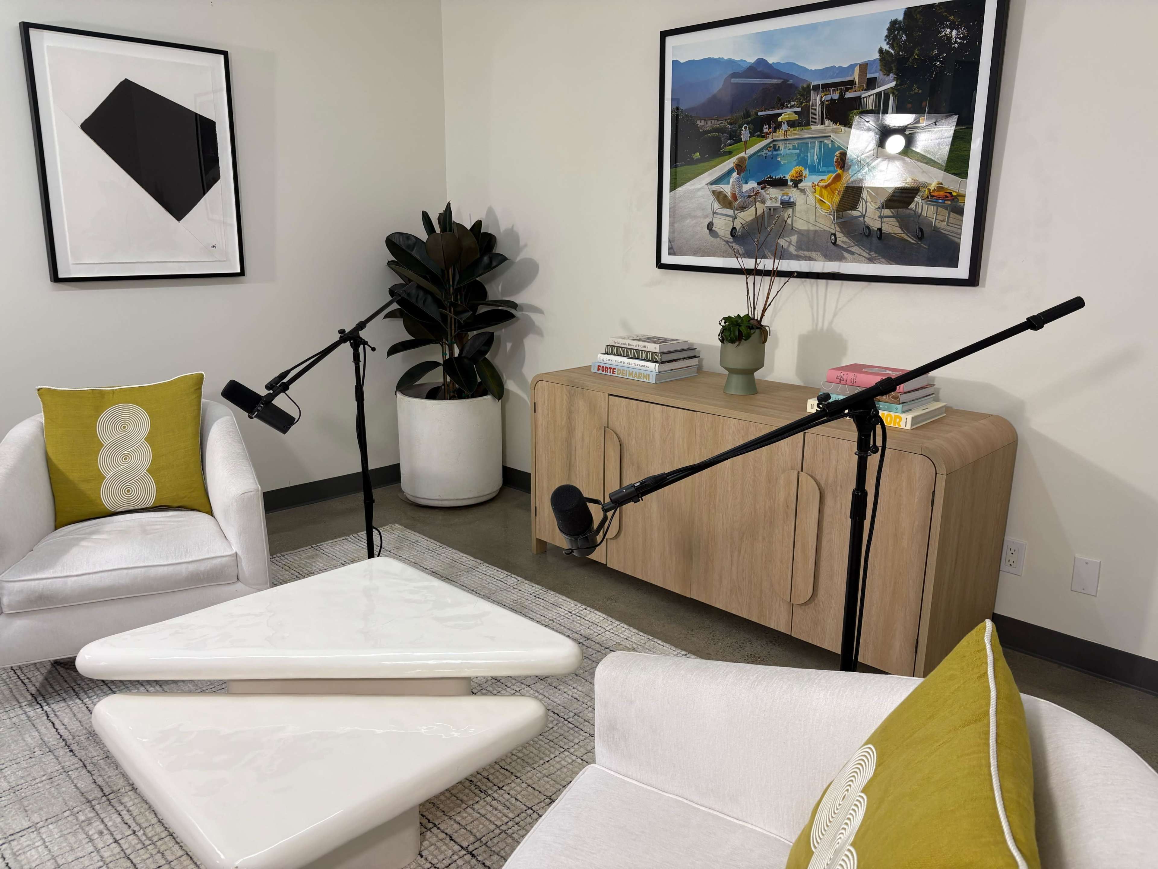 The image shows a modern interview or podcast setup featuring two armchairs, a triangular coffee table, and two microphones on stands, with artworks and a plant in the background.