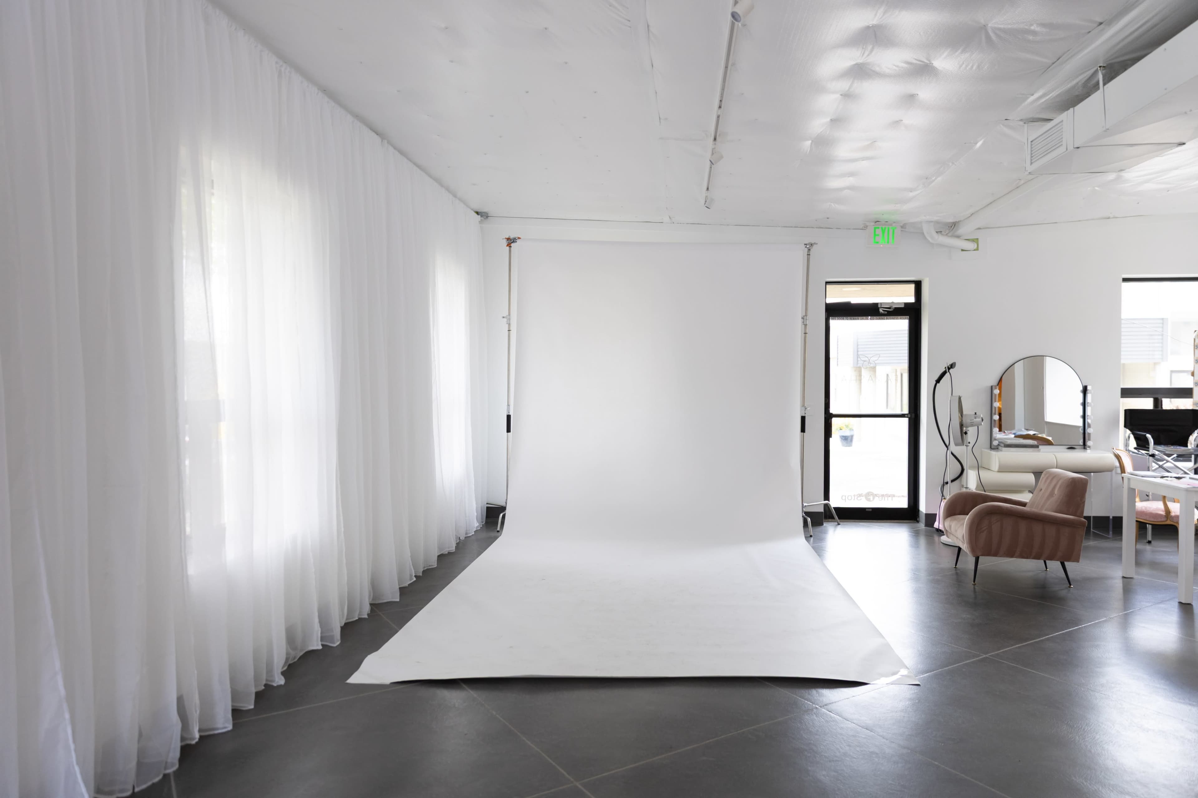 The image shows a spacious room with white drapery, a backdrop stand with a plain white backdrop, and various pieces of modern furniture.