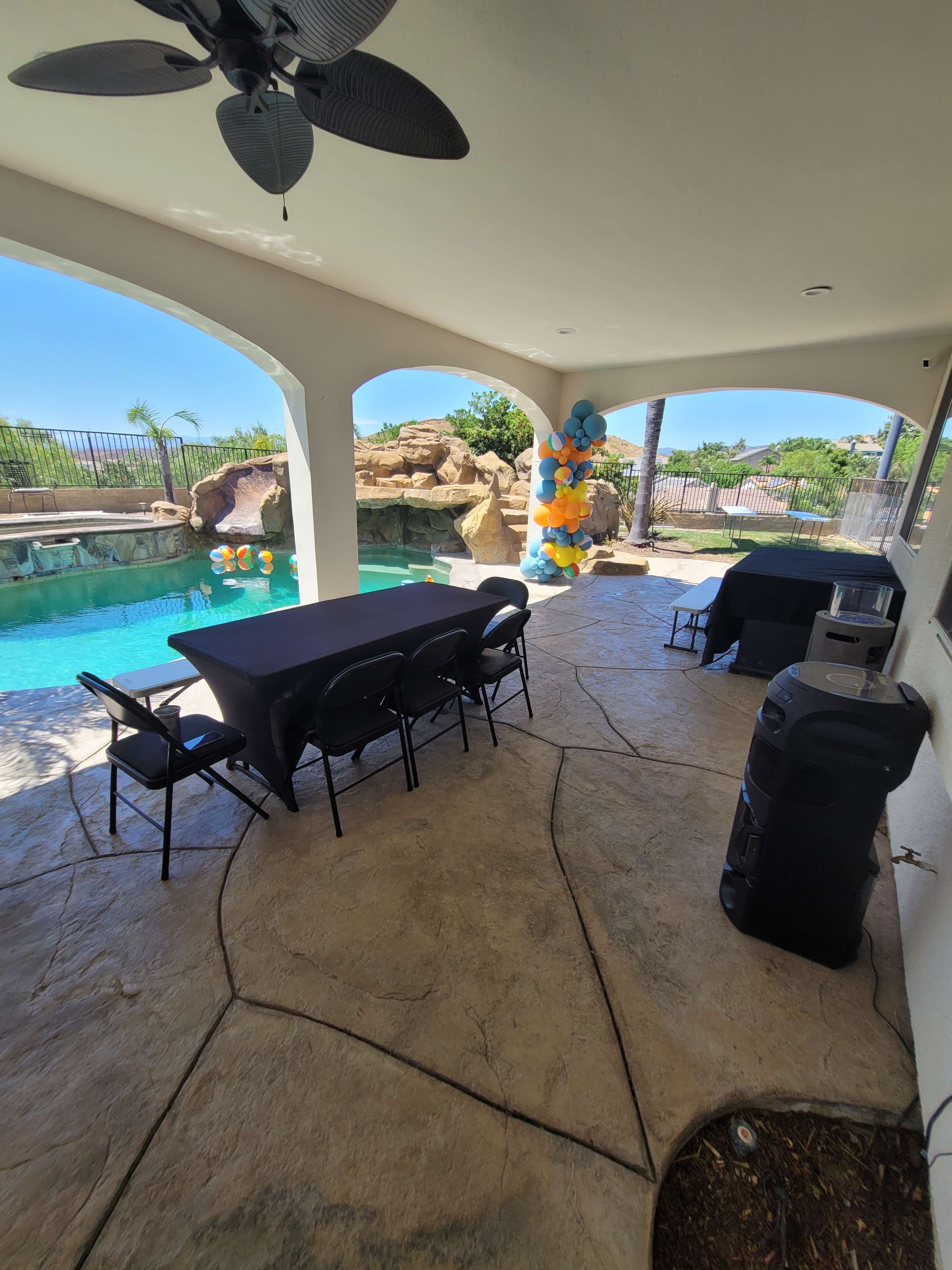 The image shows a covered patio area with black tables and chairs, a speaker, and blue and orange balloons near a swimming pool.