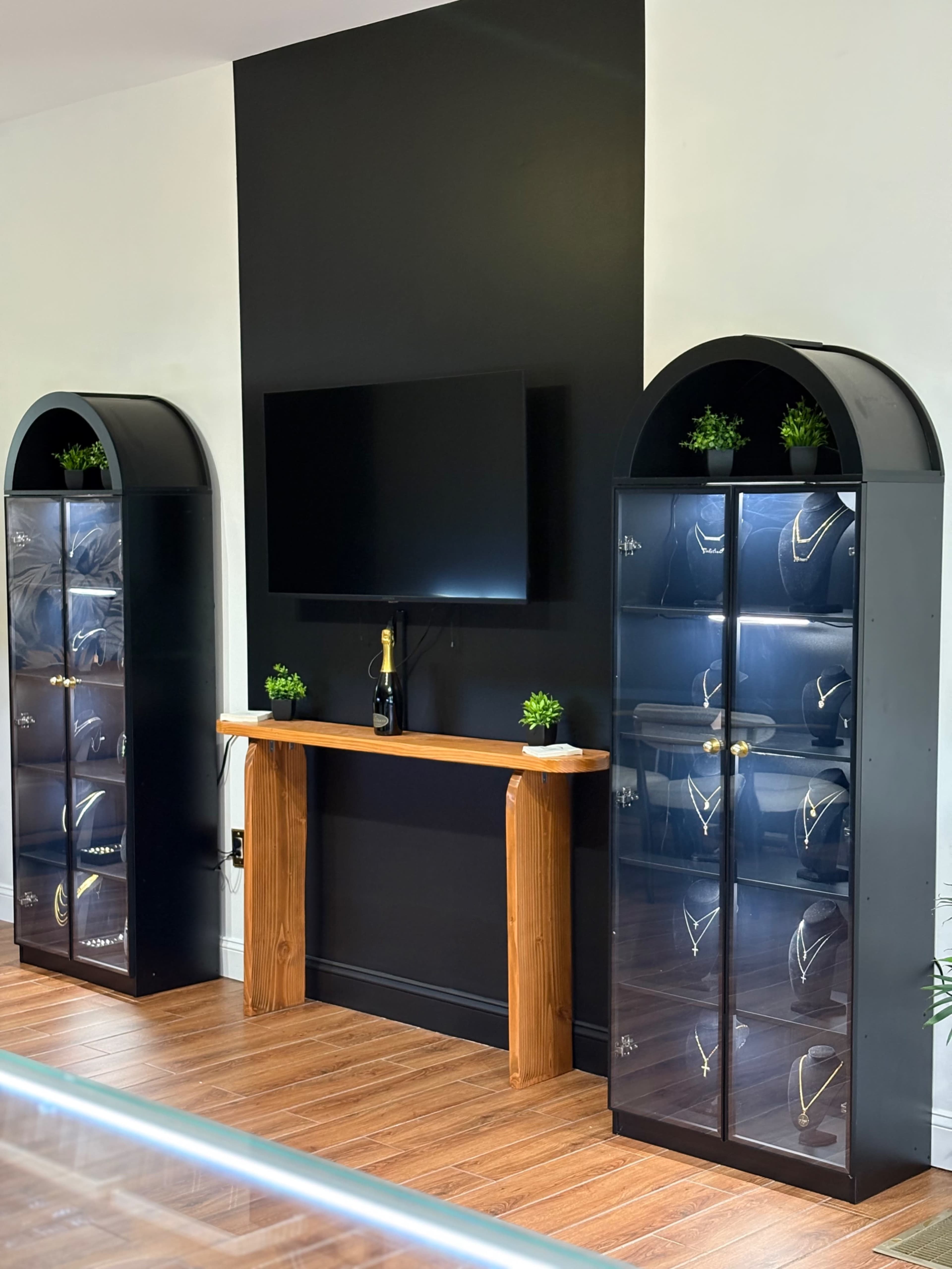 The image shows a modern display area featuring two glass display cases containing jewelry, a wooden table in the center, and a wall-mounted television above it.