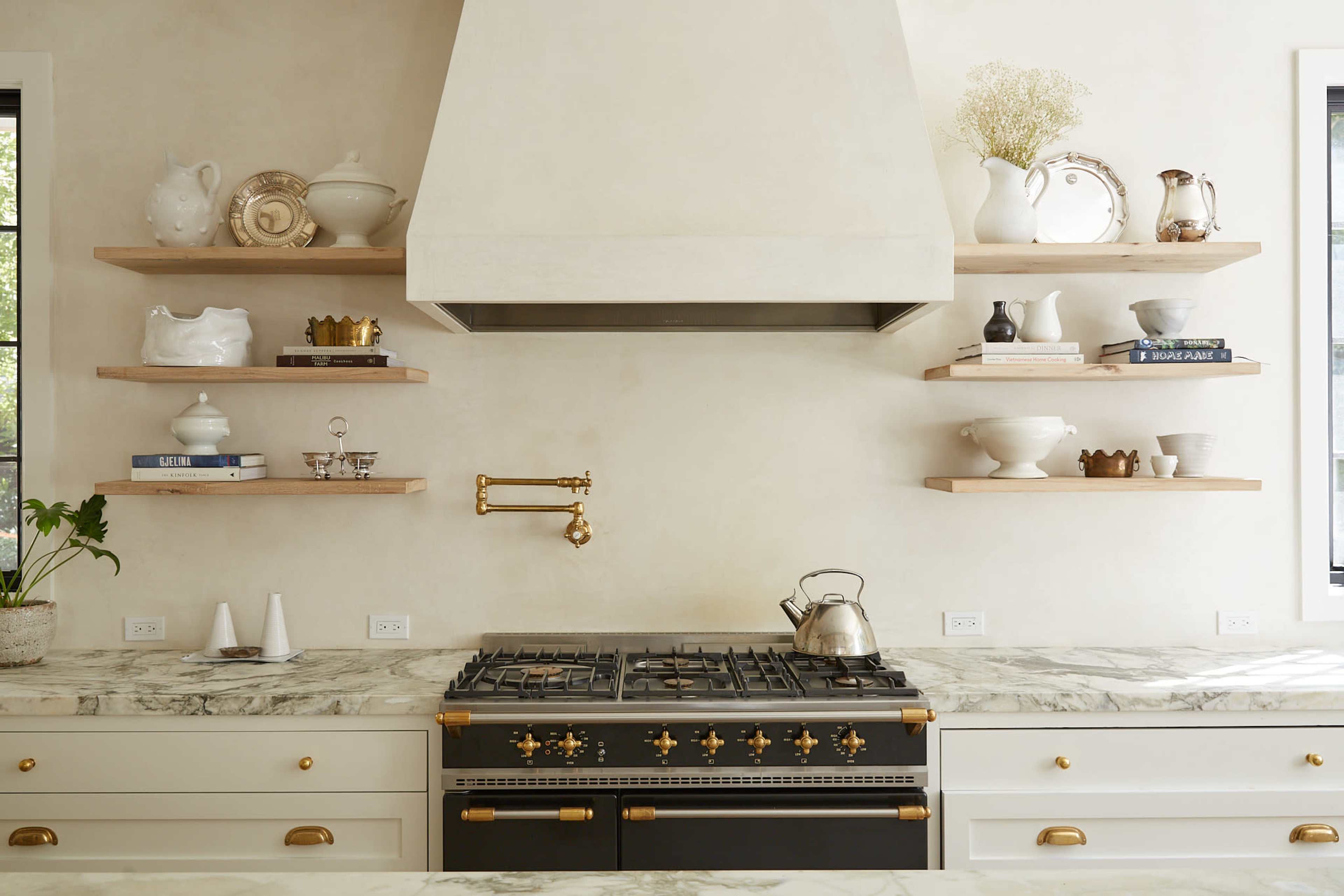 A modern kitchen with a marble countertop, a black and gold stove, and open wooden shelves displaying white dishware.