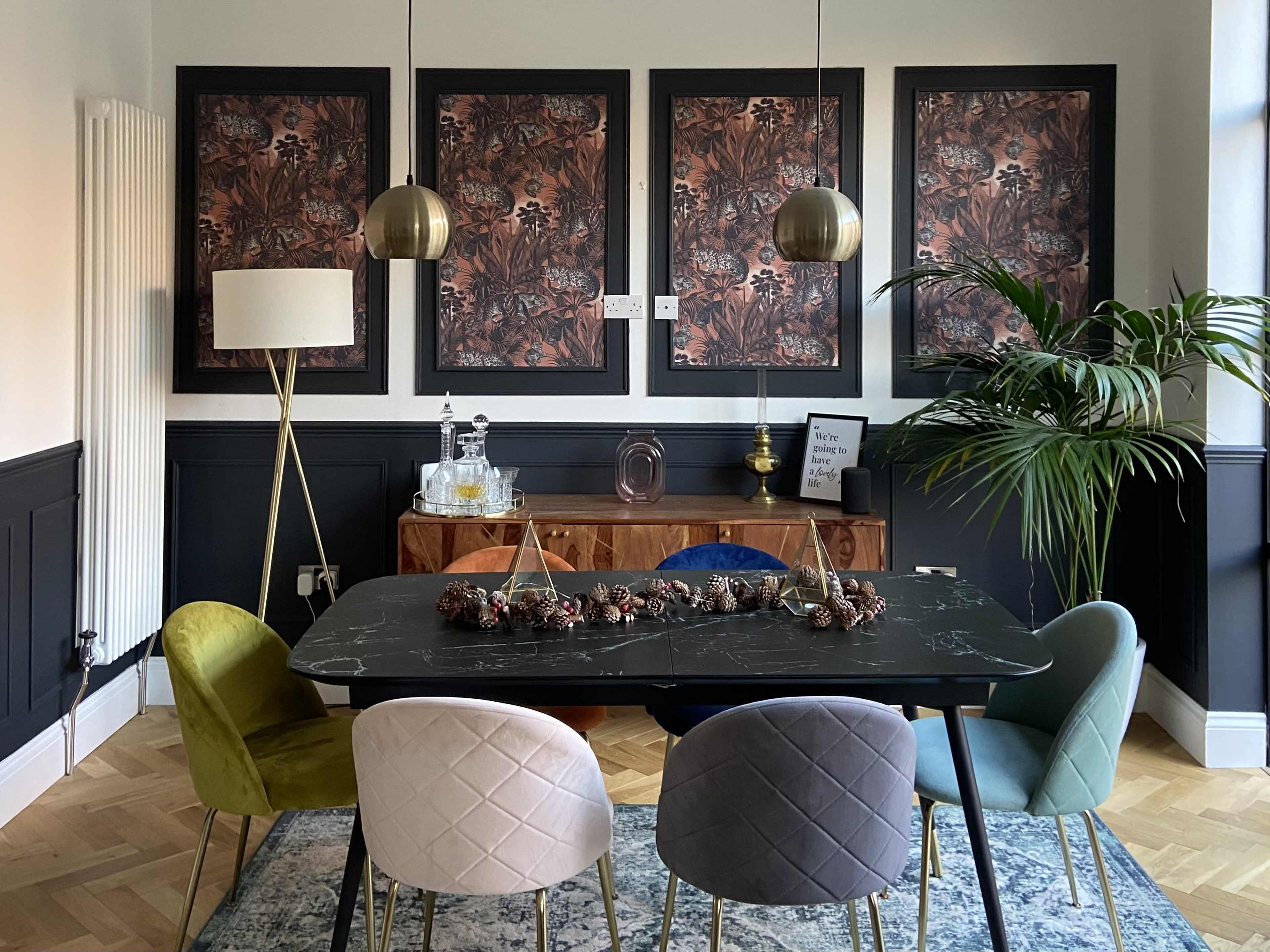 A dining area features a black table surrounded by colorful chairs, with three botanical prints on the wall and a large tropical plant in the corner.