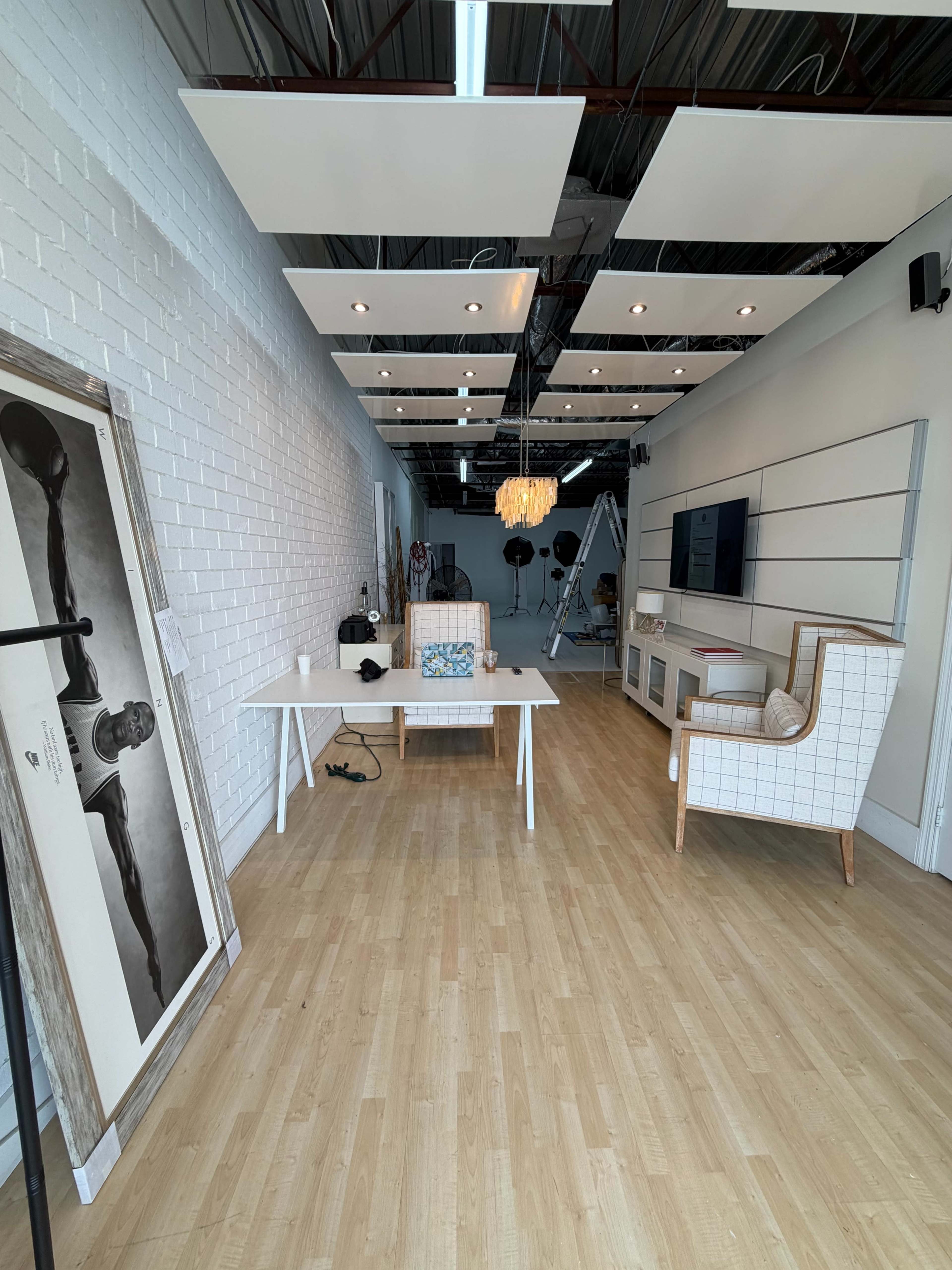 The image shows a modern, minimalistic interior of a studio space with wooden flooring, white brick walls, a desk at the center, and a large wall-mounted screen.