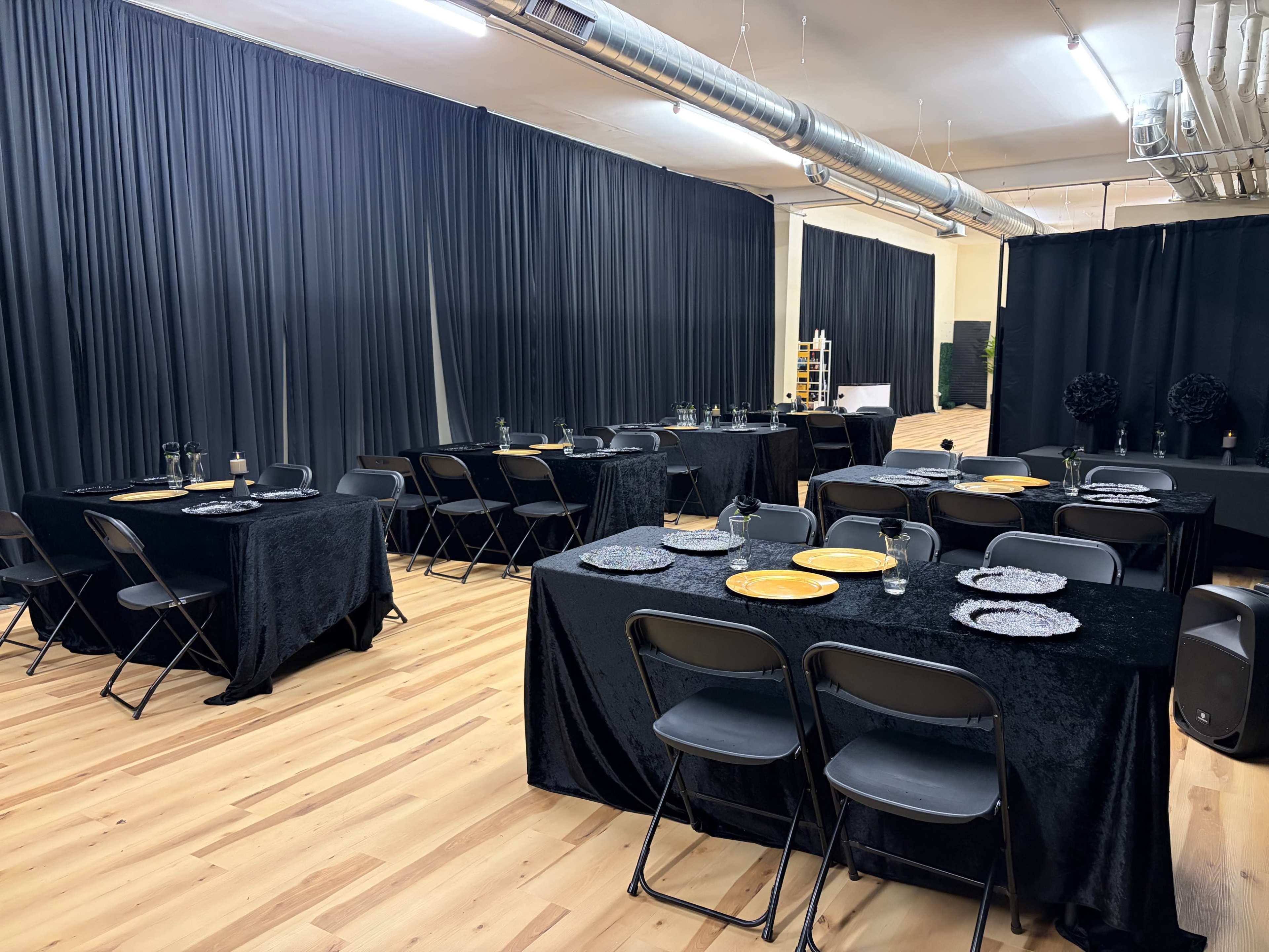 The image shows a spacious room set up for a formal dining event, with several black tablecloth-covered tables arranged neatly, each containing plates, glasses, and utensils.