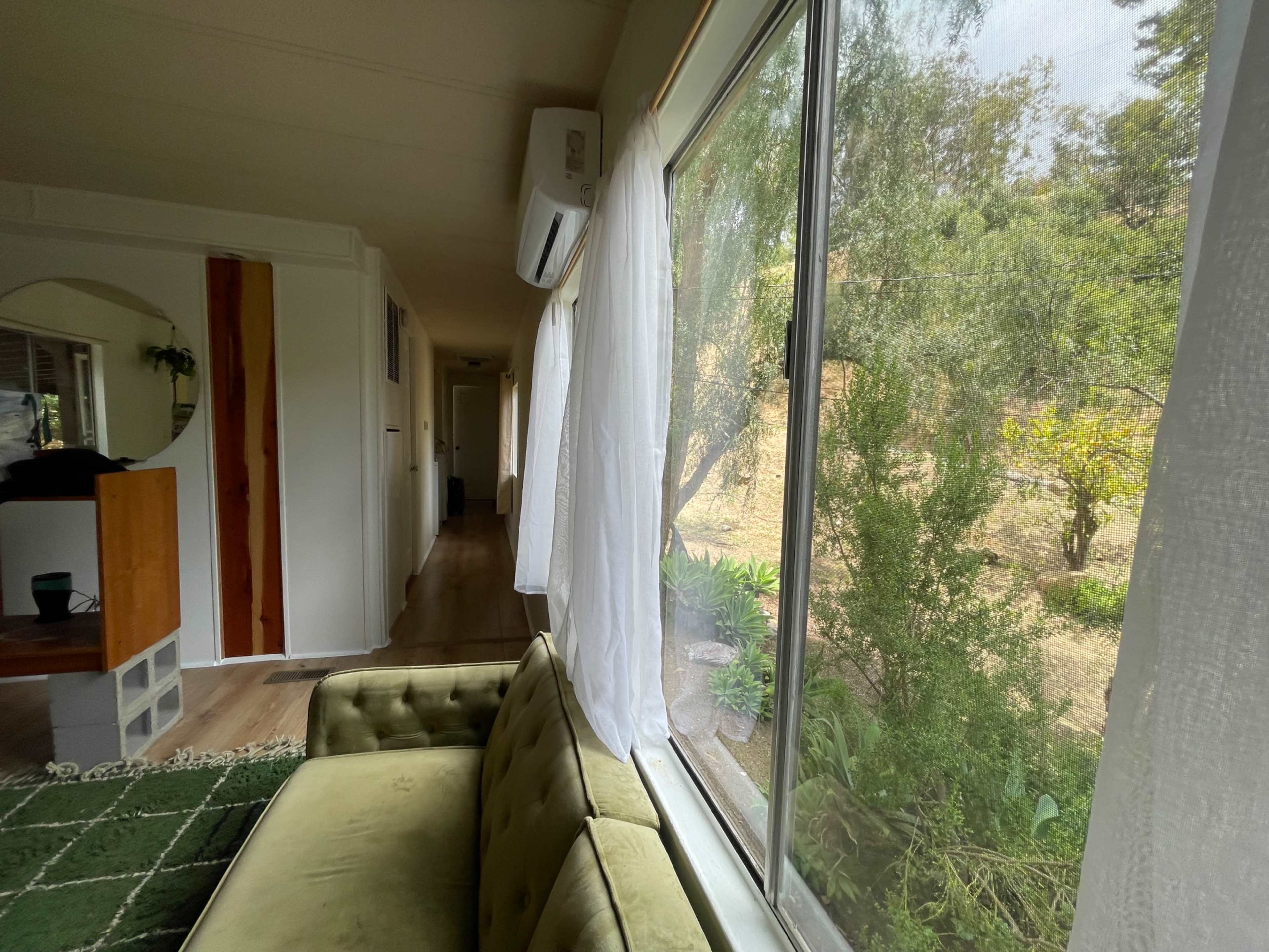 A green velvet couch sits beside a large window with sheer white curtains, overlooking a garden area.