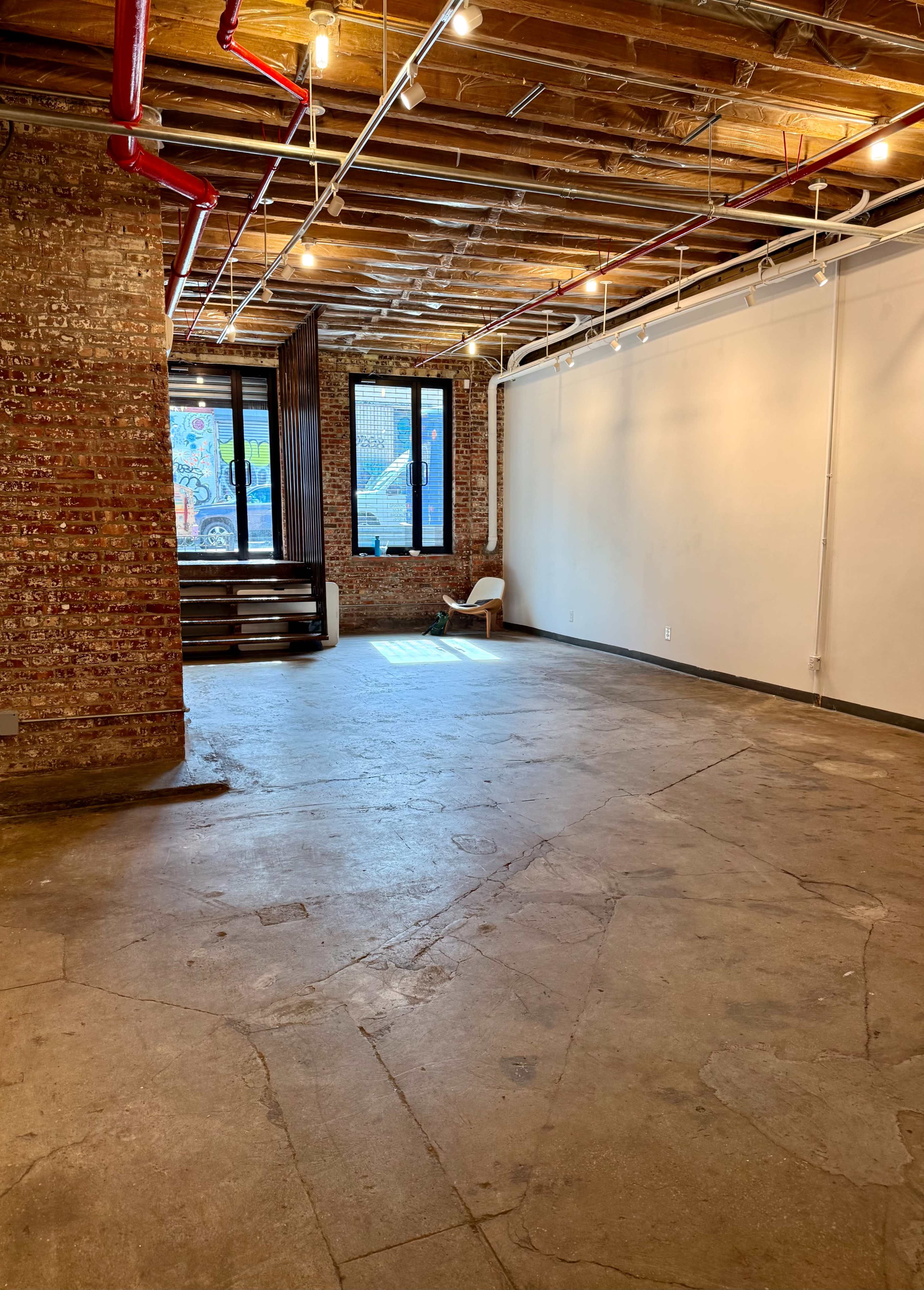 The image shows an empty room with exposed brick walls, wooden beams, and concrete flooring, featuring large windows that allow natural light to enter.