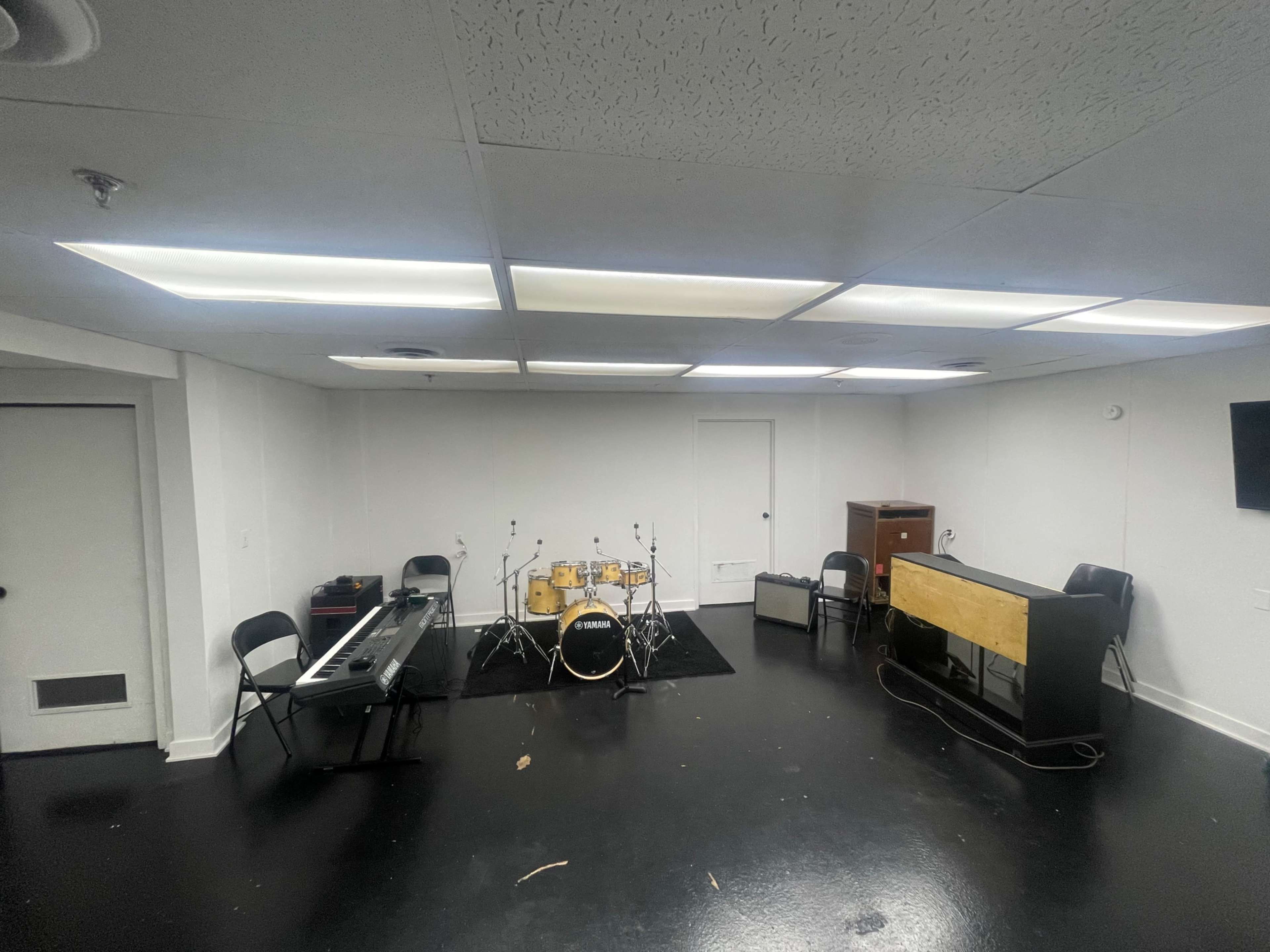 The image shows a music rehearsal room with a drum set, keyboard, amplifier, and several folding chairs arranged around the space.