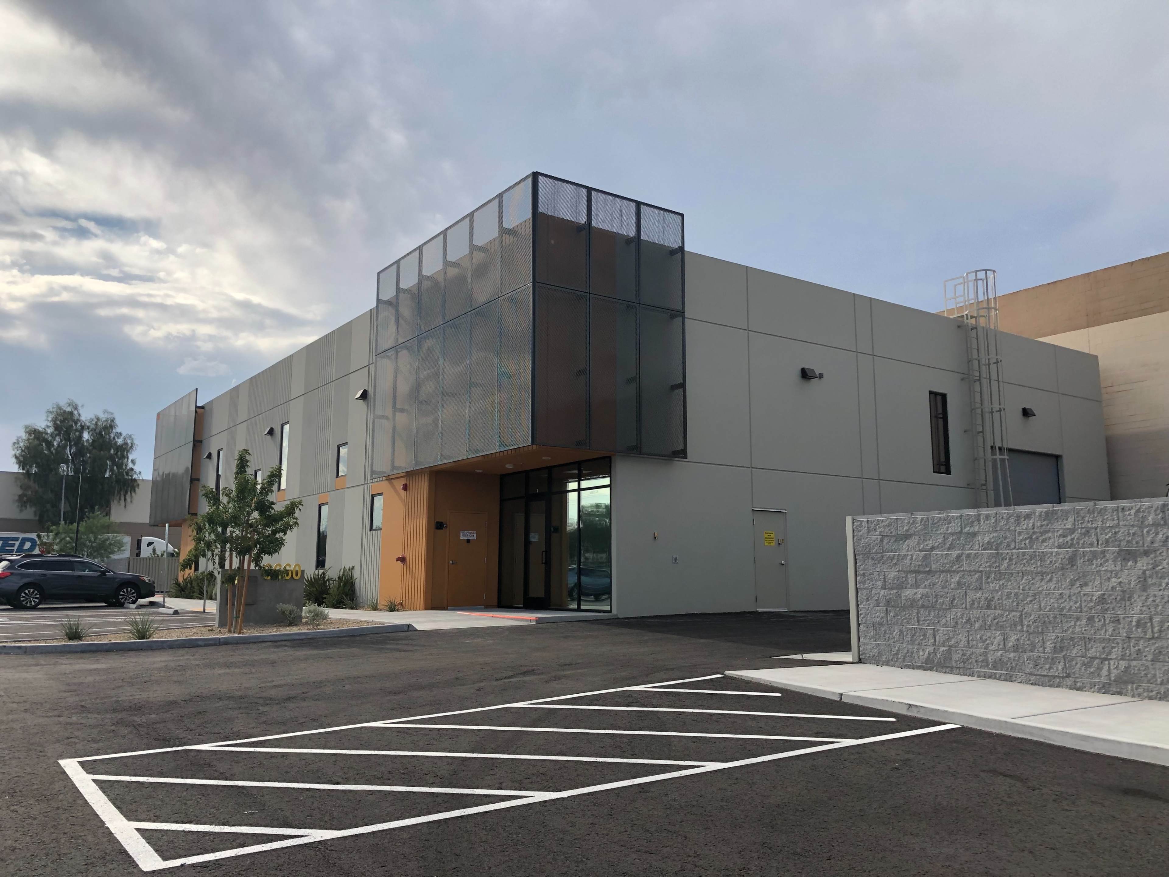 The image shows a modern, two-story building with large windows and a gray exterior, situated next to a paved parking lot with marked spaces.