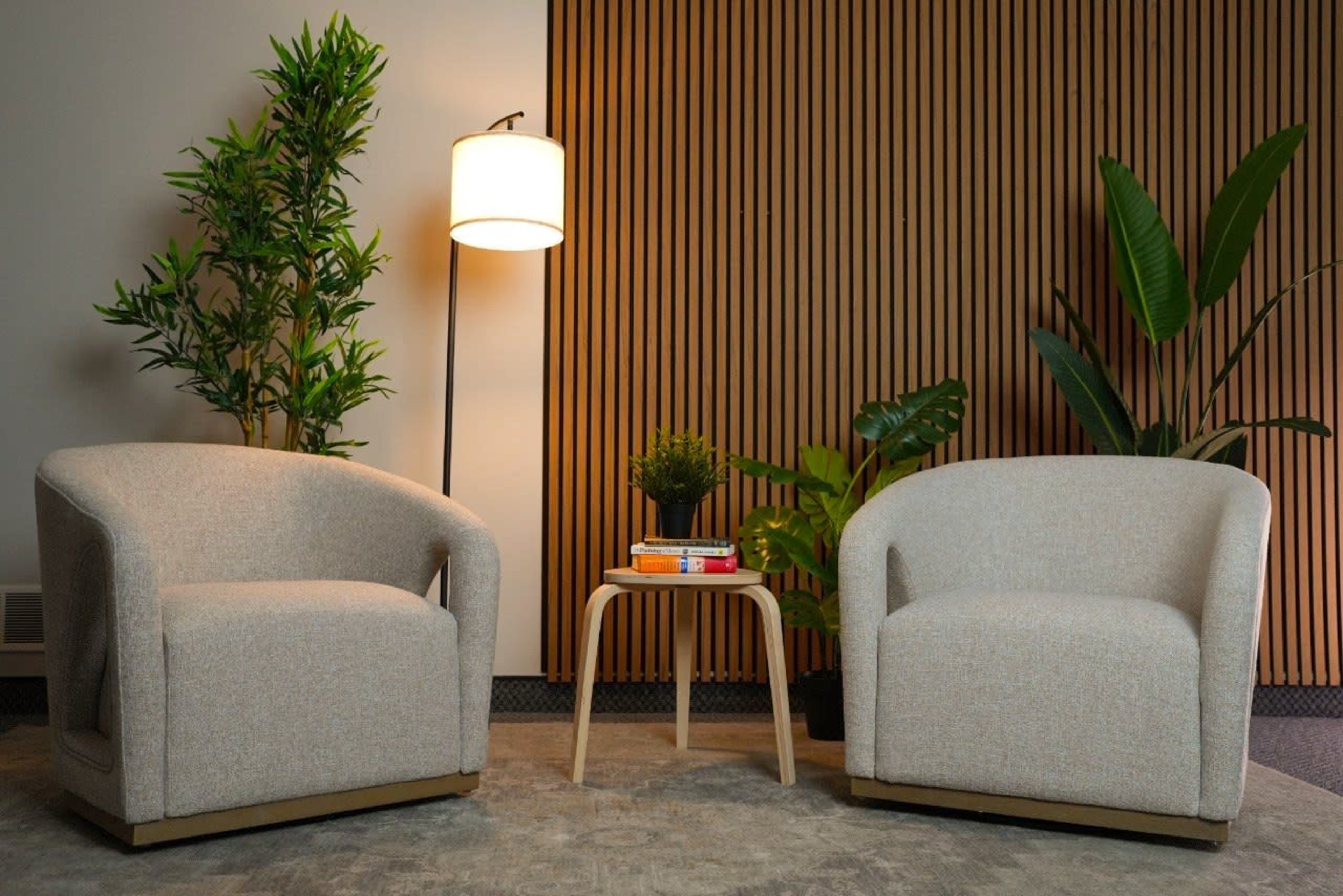 The image shows two textured armchairs positioned opposite each other with a small wooden table between them, surrounded by indoor plants and a lamp.