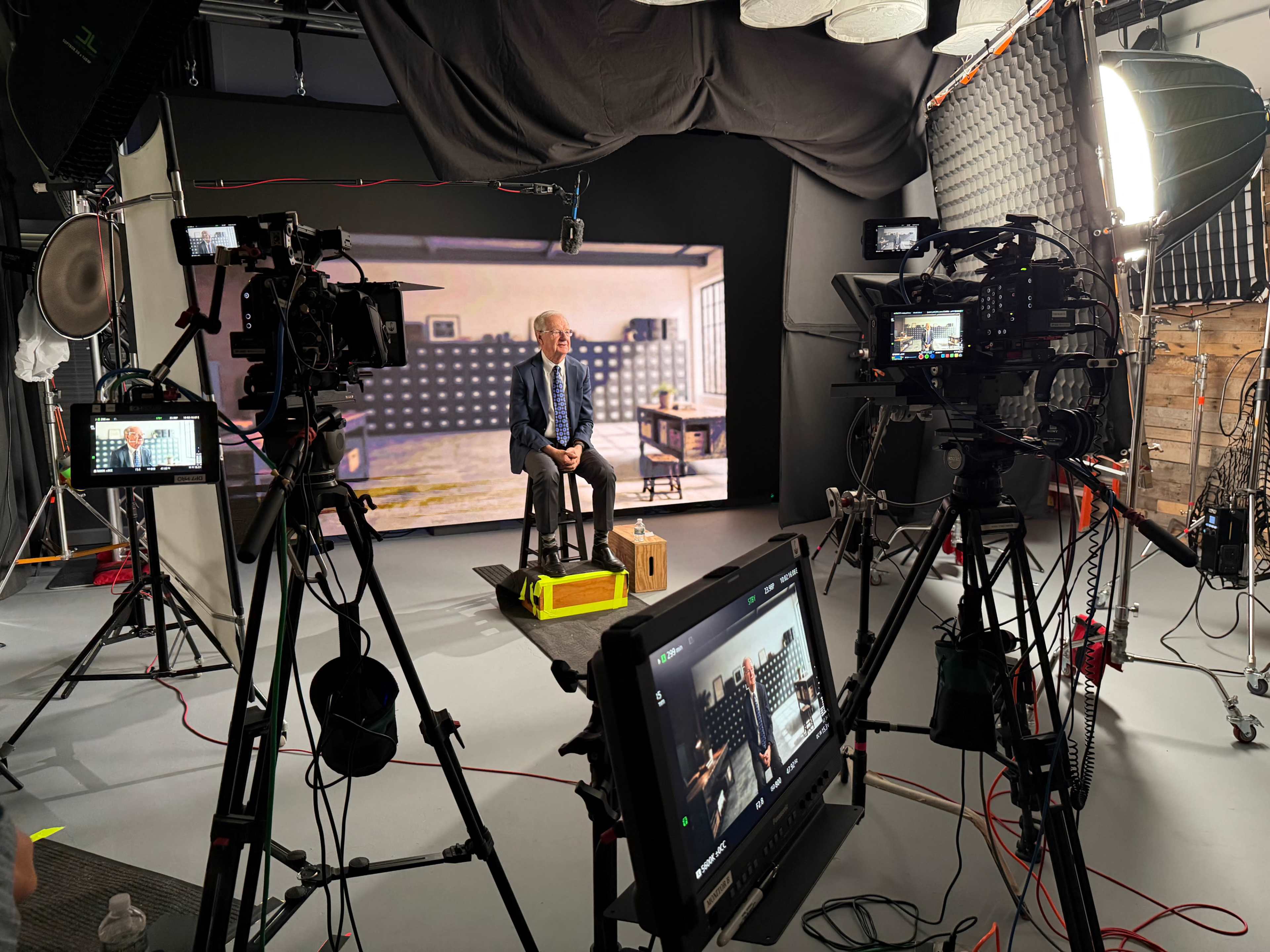 A studio setup features multiple cameras and lights focused on a man seated on a stool in front of a backdrop depicting an office interior.