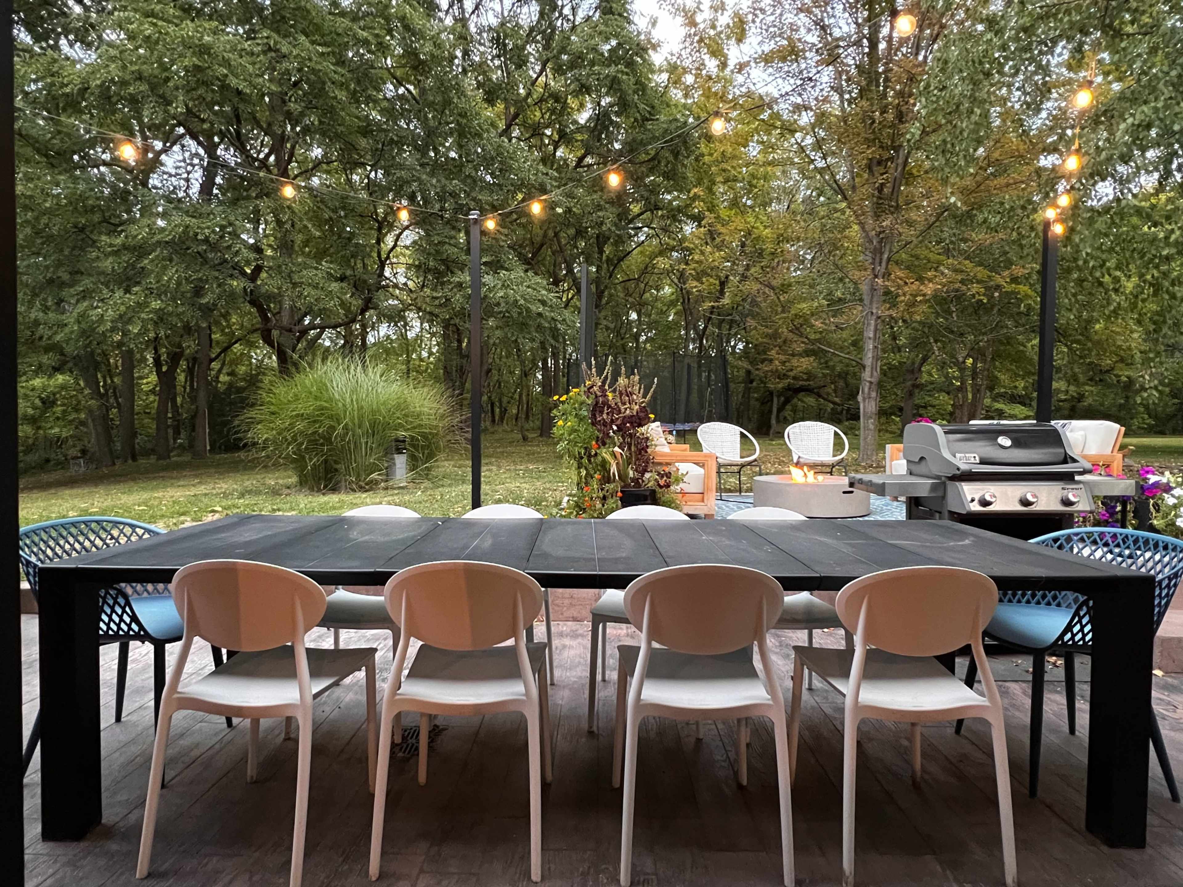 A long black dining table with white chairs is set on a patio facing a lush green yard illuminated by string lights.