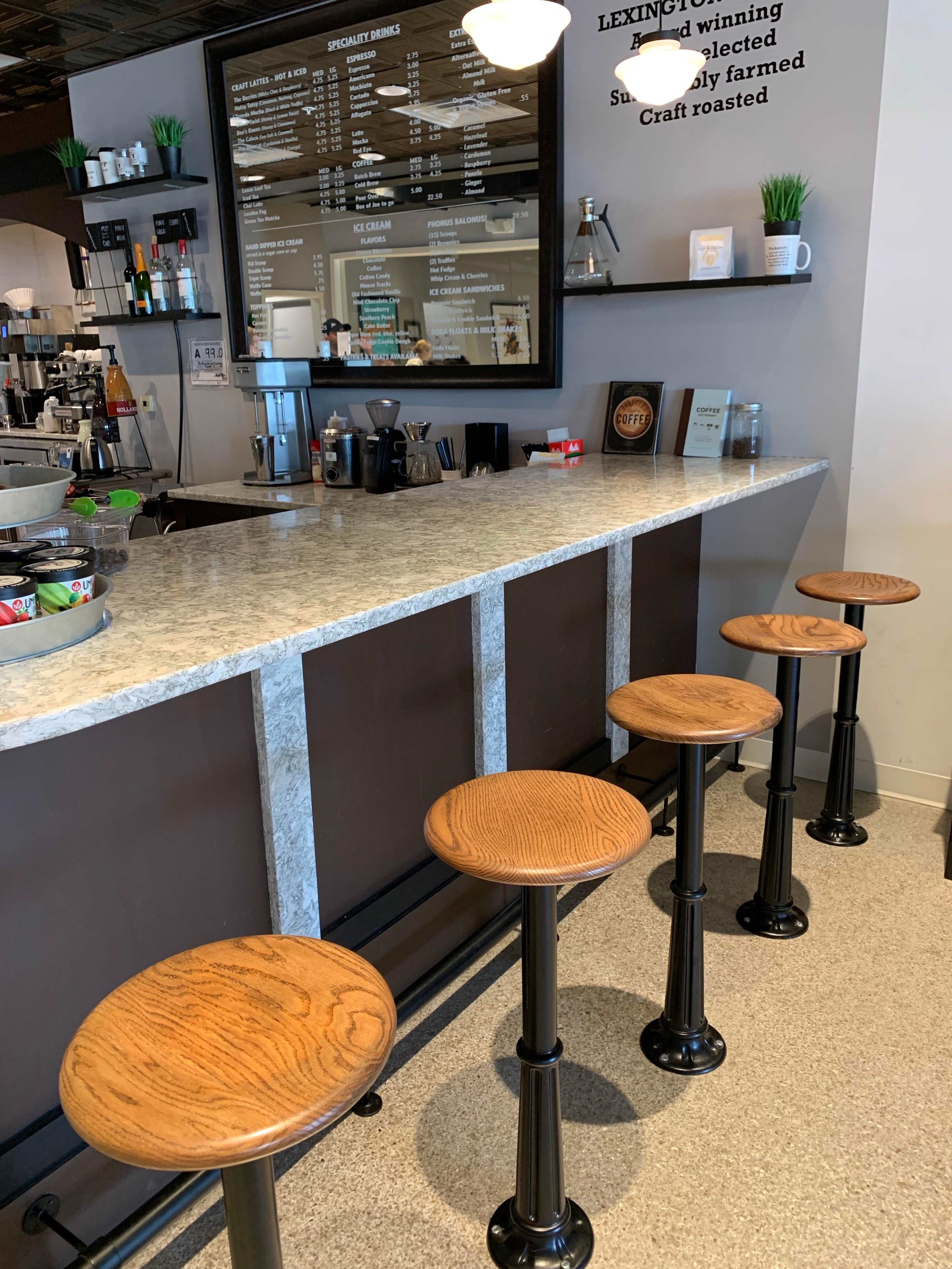 A coffee shop counter with four wooden stools arranged in front of a granite surface, featuring a menu board and various coffee-making equipment behind it.