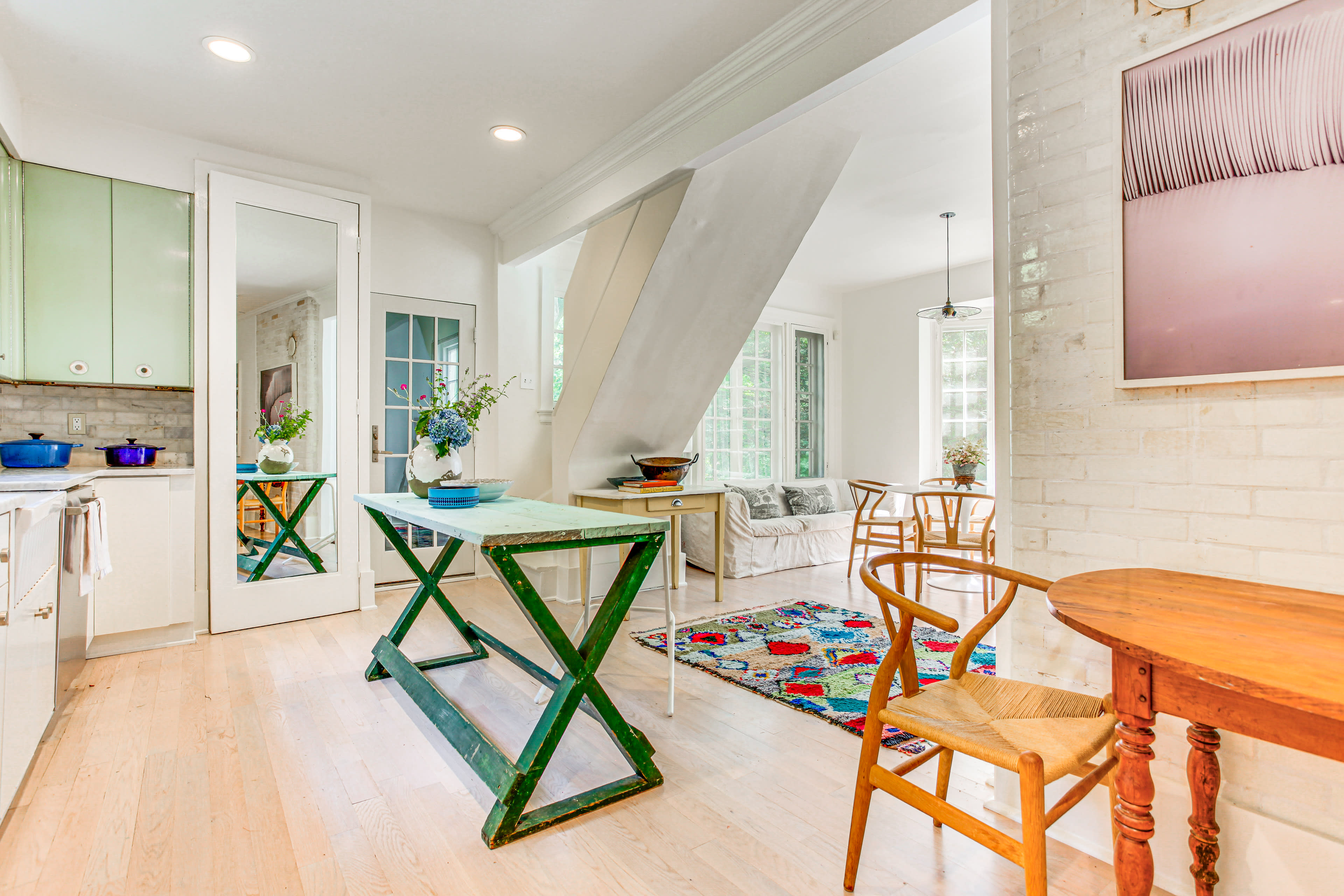 The image shows a well-lit dining area with a green table, wooden chairs, and a cozy seating space, featuring large windows and a vibrant area rug.