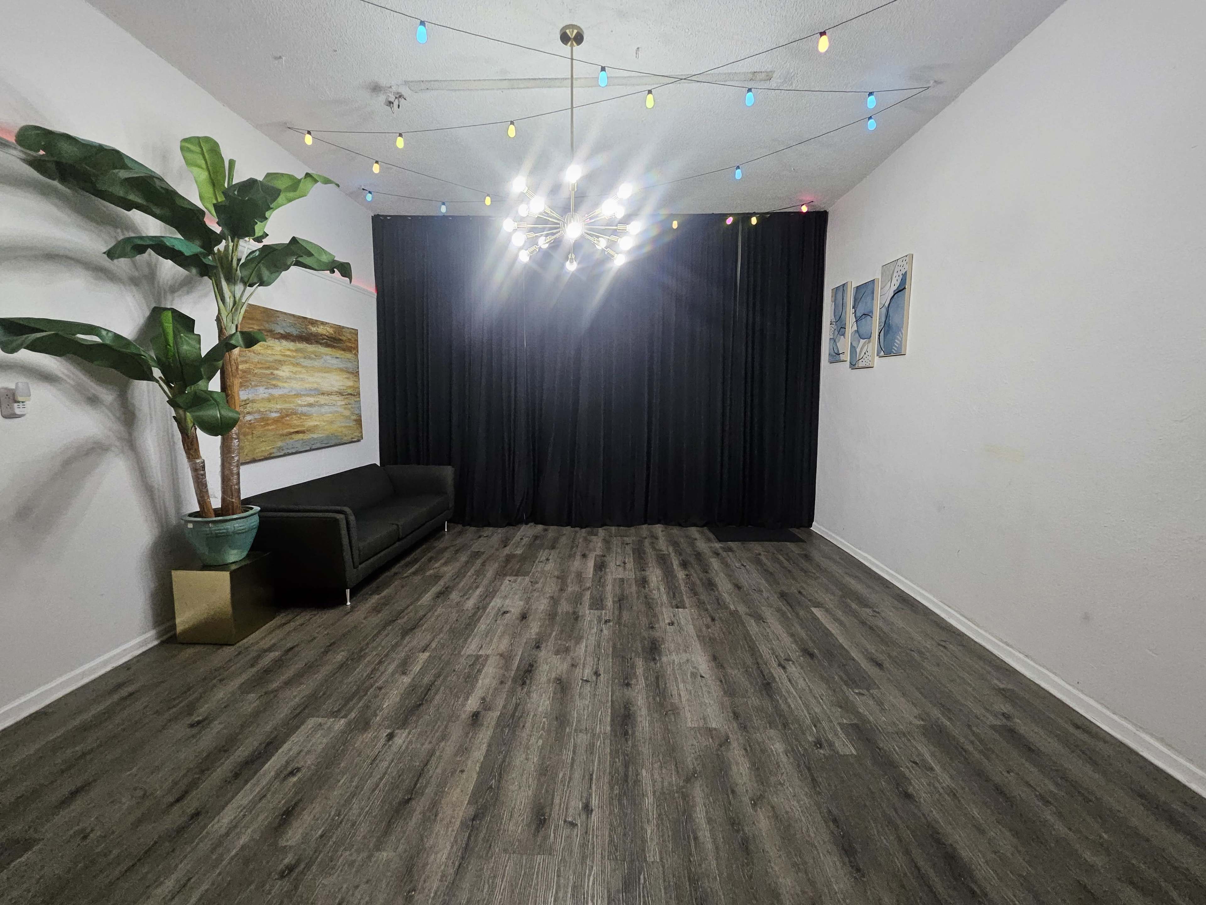 The image shows a spacious room with a dark curtain backdrop, a modern sofa, colorful string lights overhead, a potted plant in the corner, and artwork on the walls.