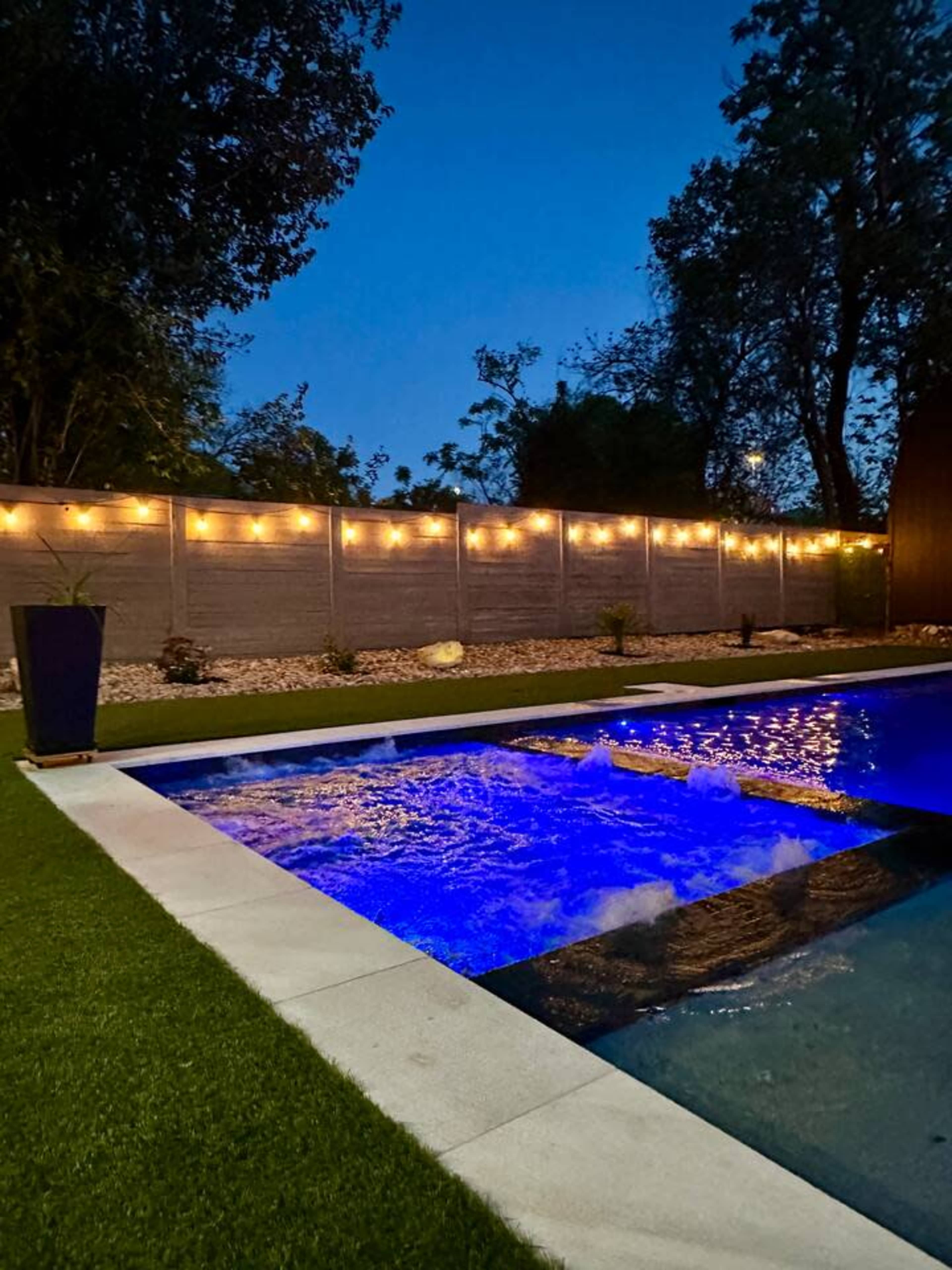 The image shows a swimming pool surrounded by a well-manicured lawn, with a decorative wooden fence lined with string lights against a twilight sky.