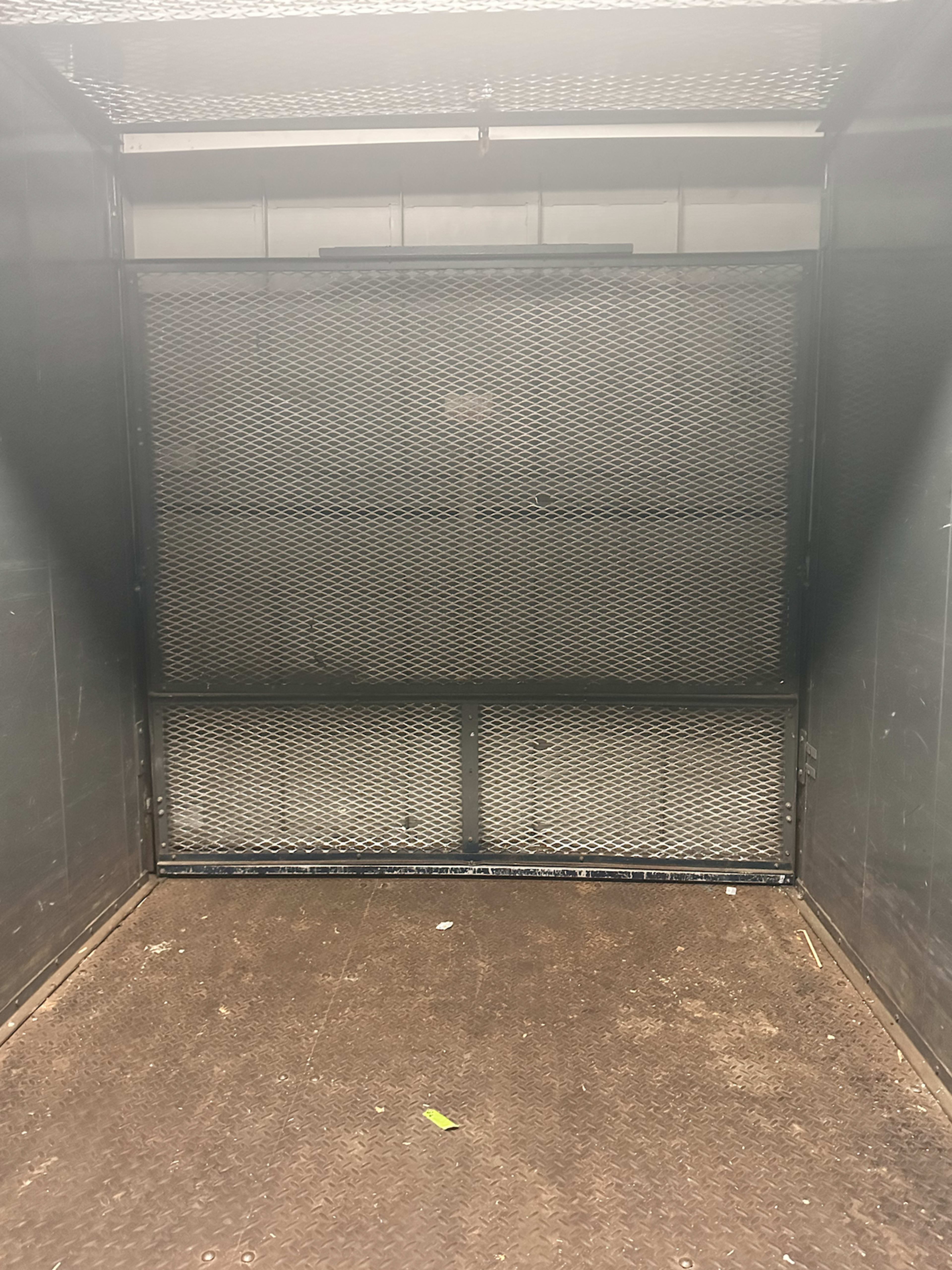 The image shows the interior of an empty metal elevator with a grated door closed at the front.