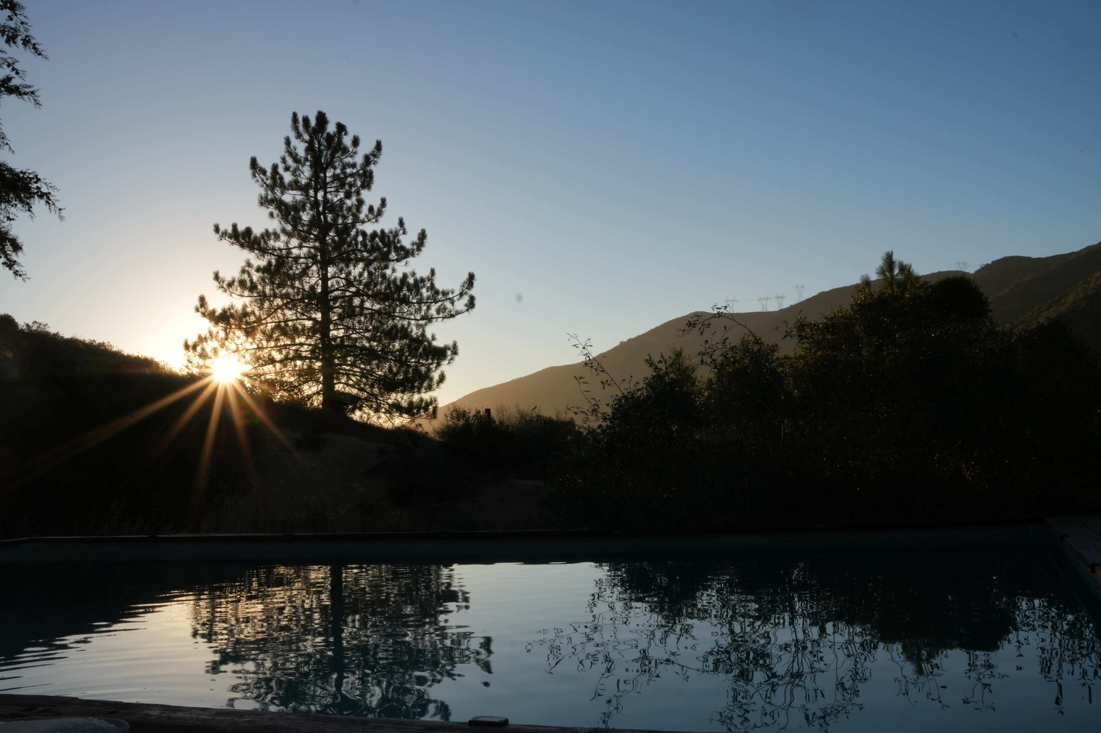 The sun rises behind a tree overlooking a still pool, with mountains silhouetted in the background.