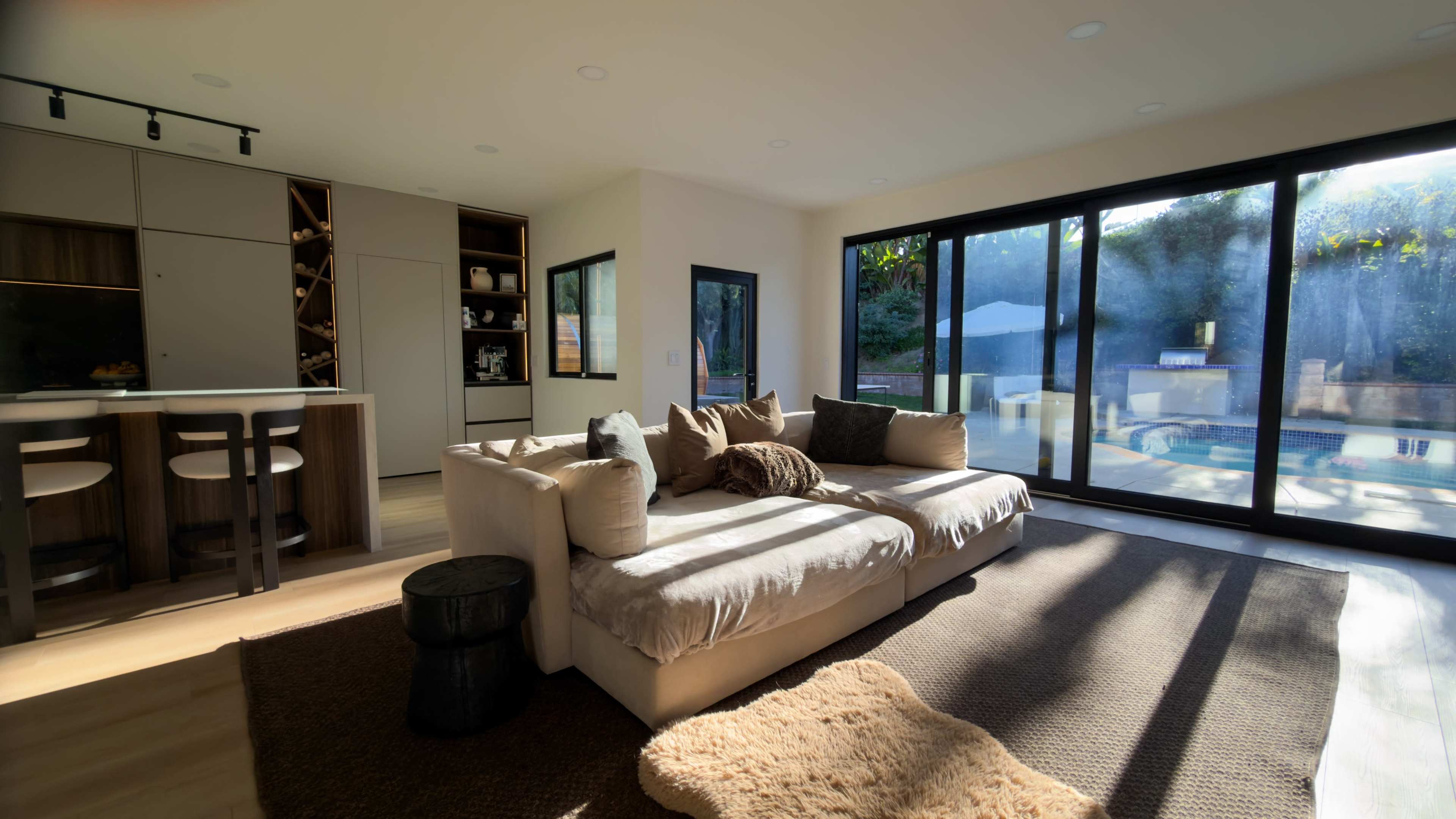 The image shows a modern living room with a light-colored sofa, a small side table, and large glass doors that open to a pool area.