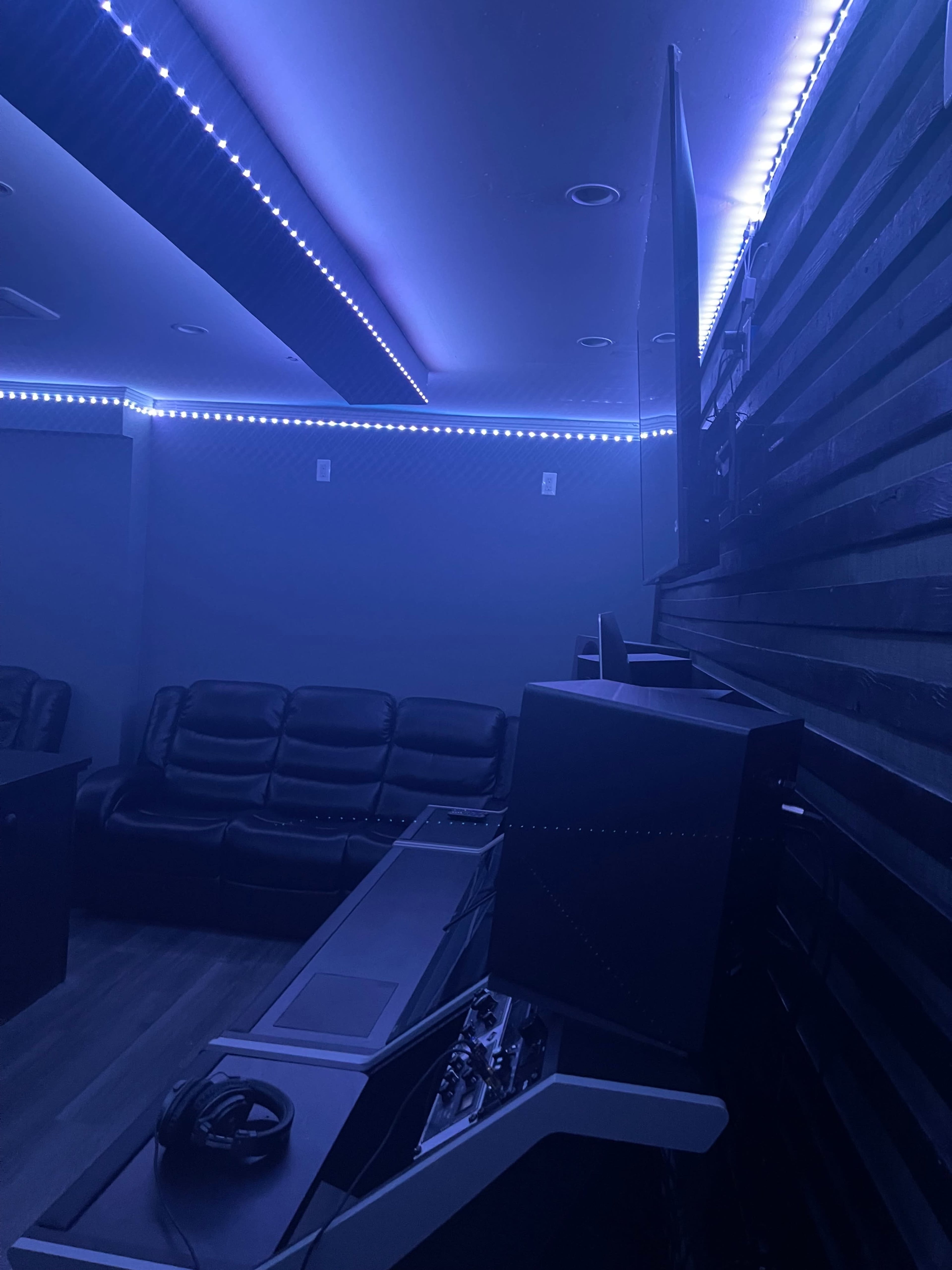 A dimly lit room with black leather couches, a computer setup, and blue LED accent lighting along the walls.