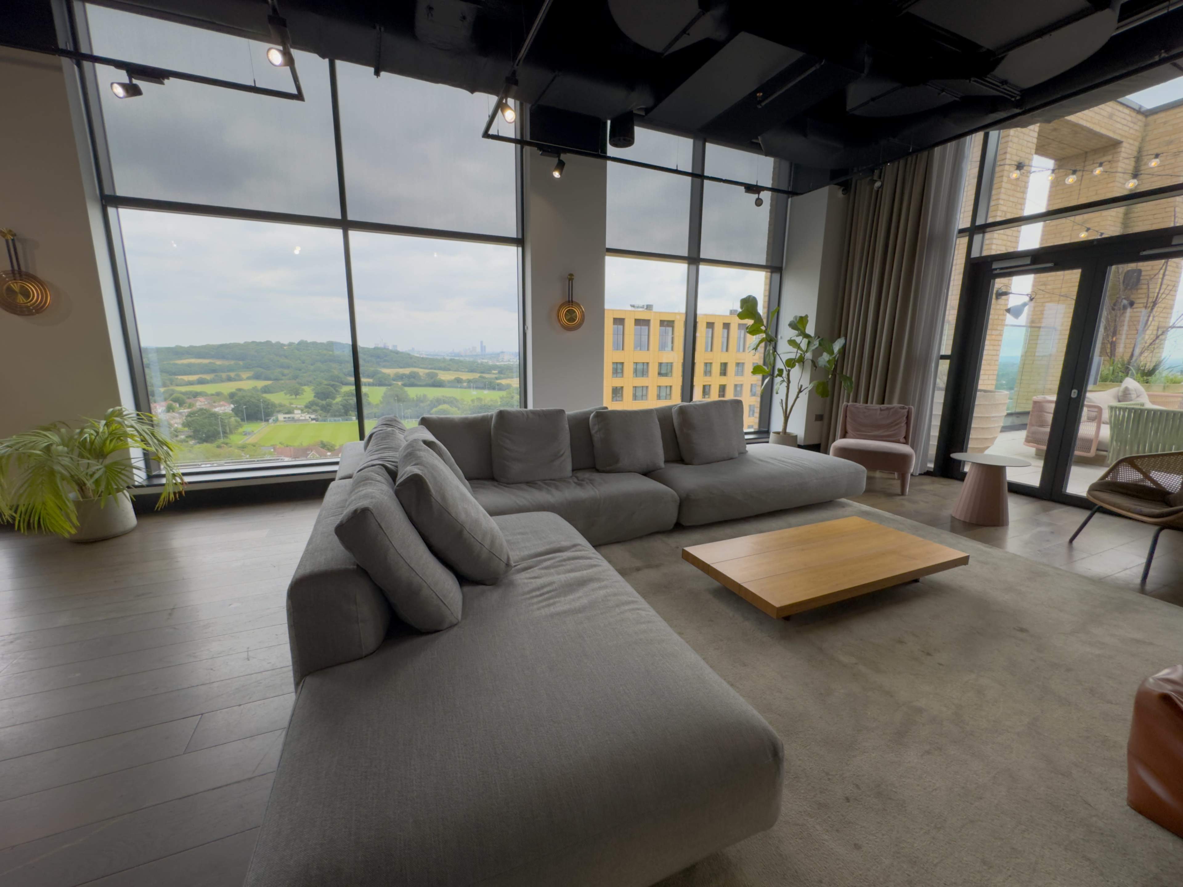 A modern living room features a spacious gray sectional sofa, a wooden coffee table, and large windows offering a view of a green landscape and buildings in the distance.