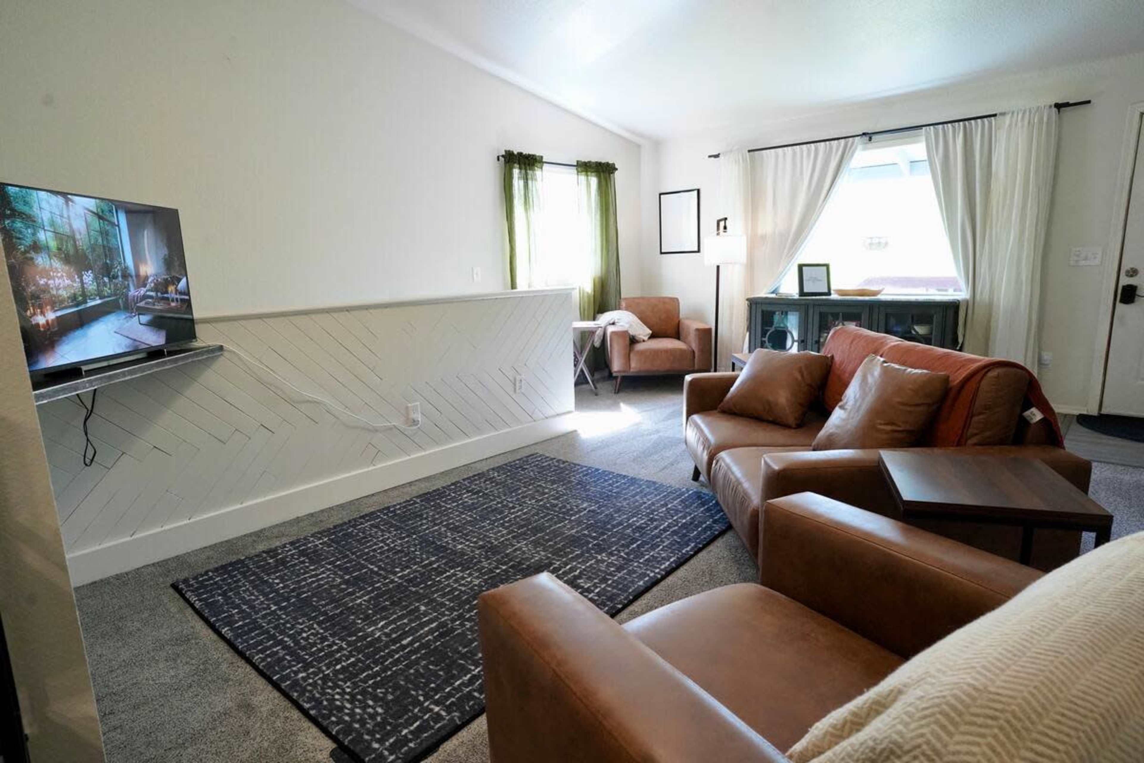 The image shows a living room with two brown leather chairs, a dark rug, a television mounted on the wall, and sunlight streaming through a window.