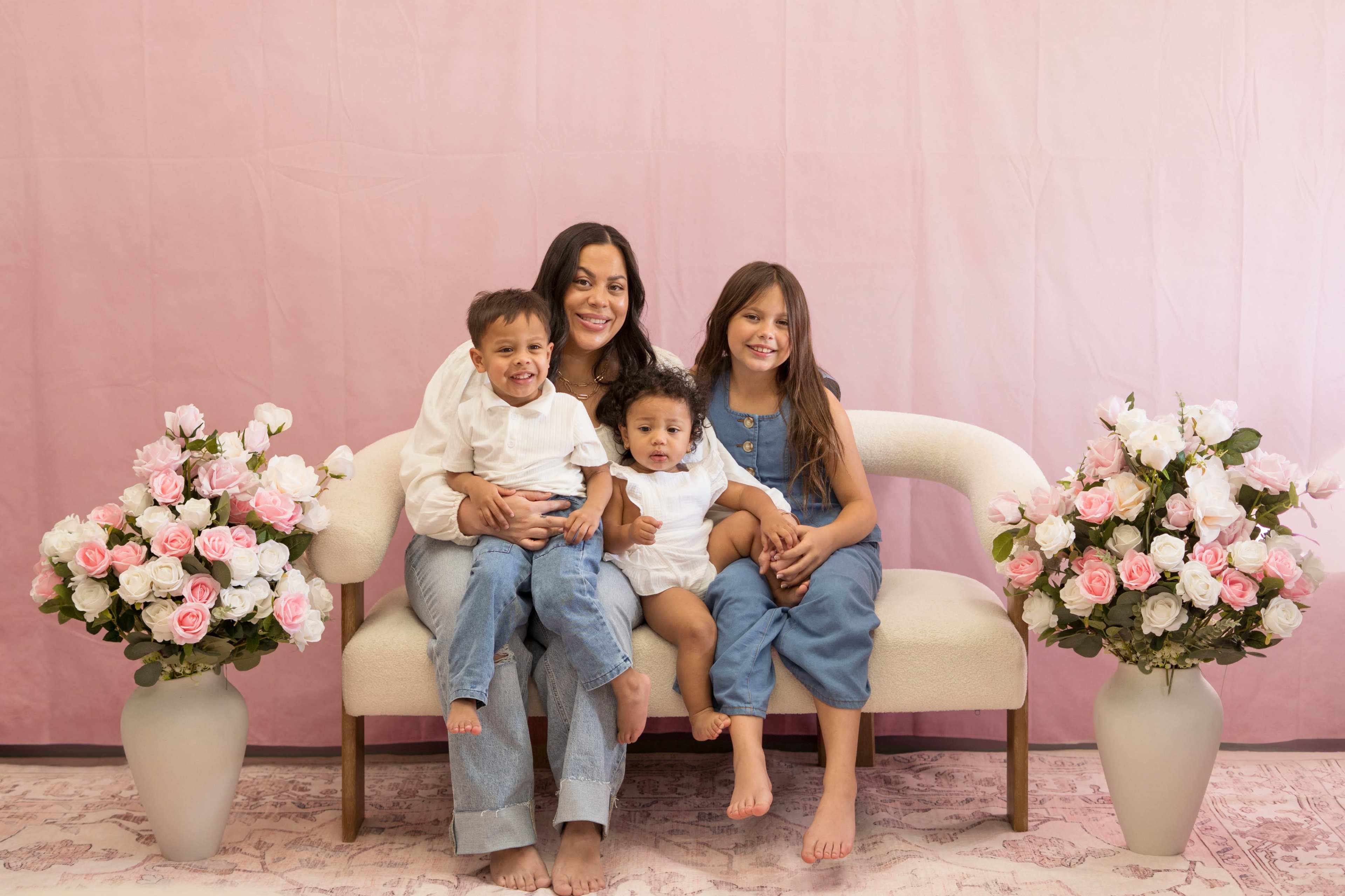 A woman sits on a light-colored couch with two young children on her lap and one beside her, with pink flower arrangements on either side and a soft pink backdrop.