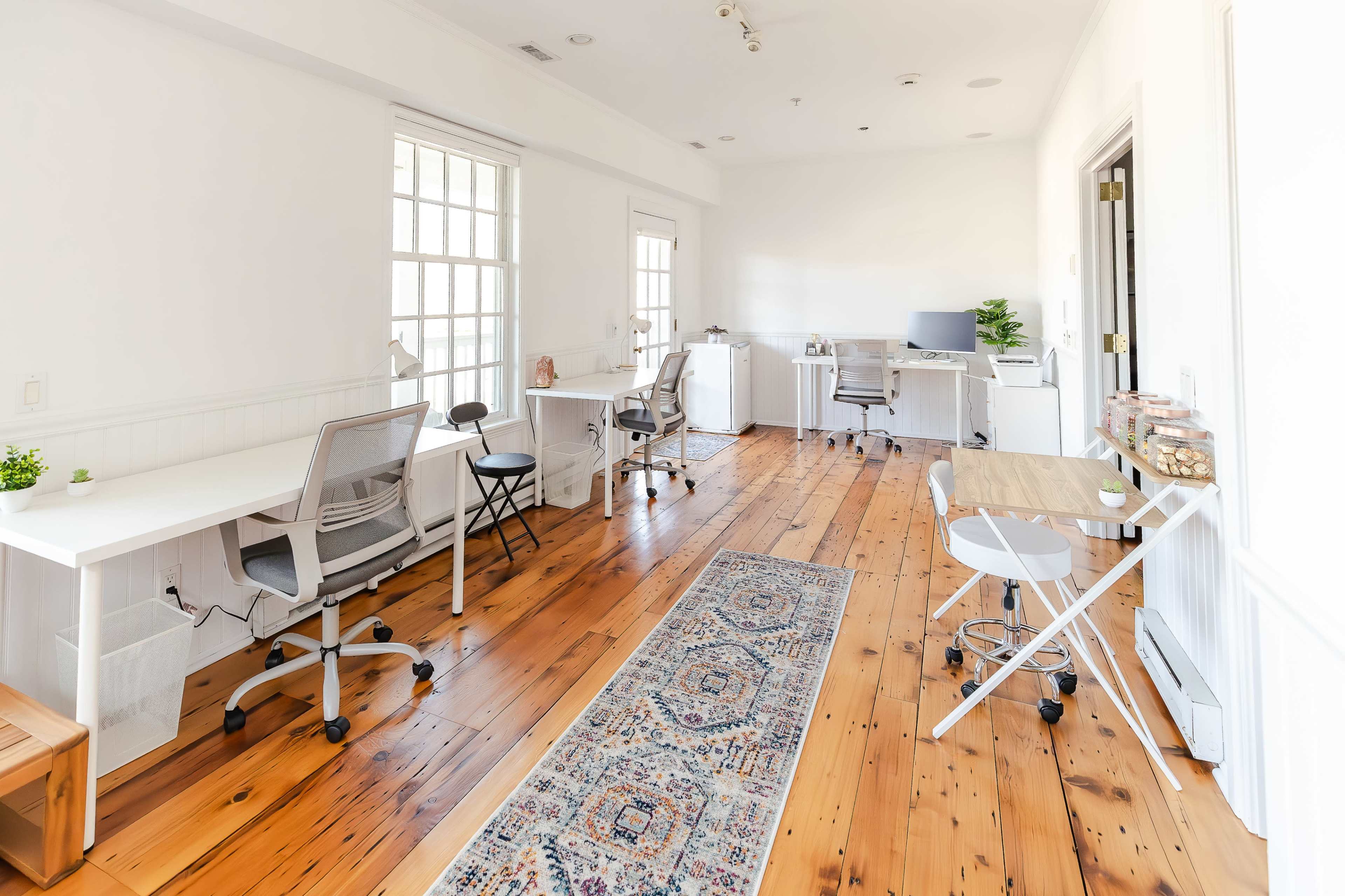 A bright, spacious office with wooden flooring, three workstations, and large windows allowing natural light to fill the room.