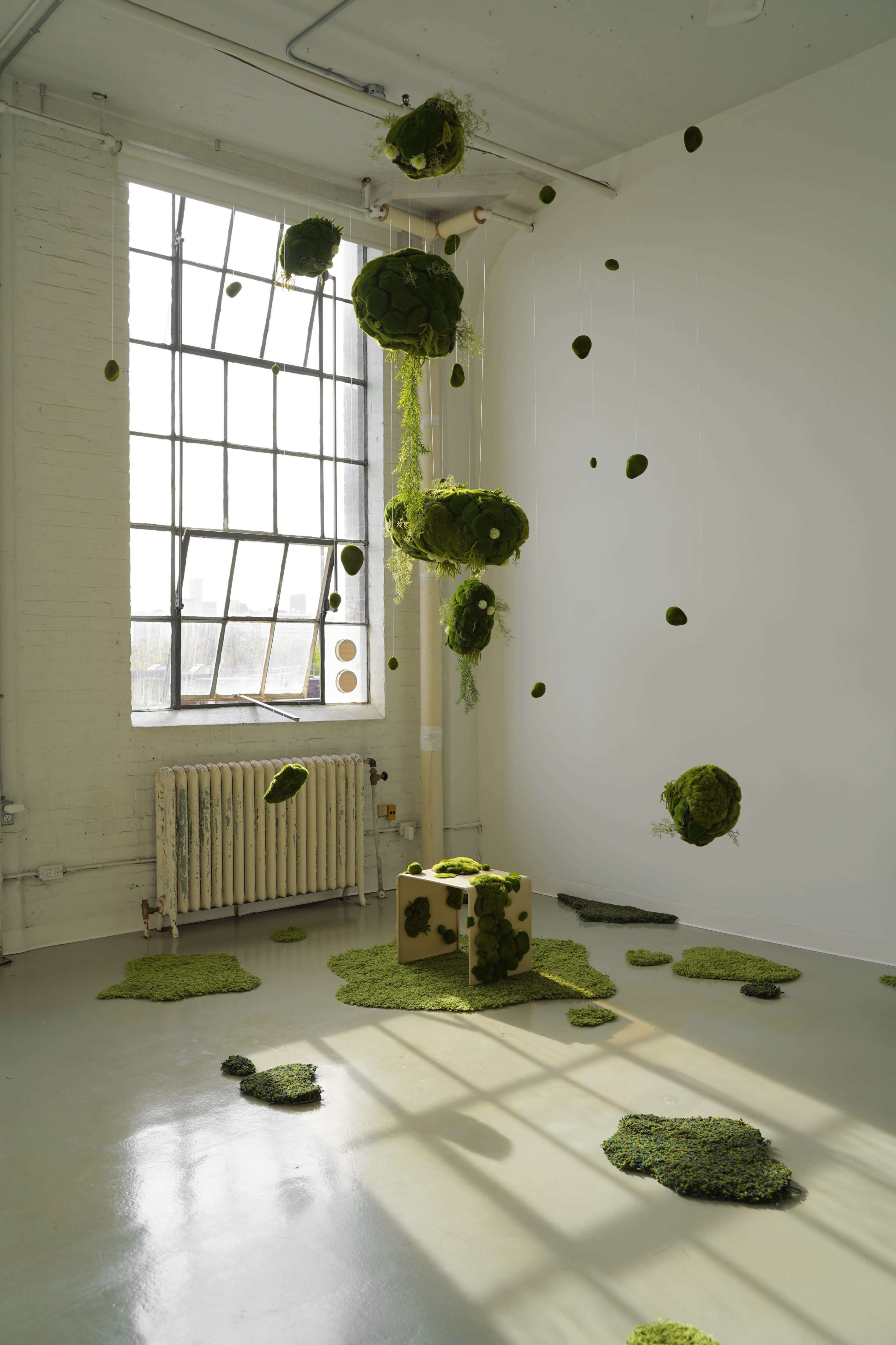 The image shows a contemporary art installation featuring green moss-like forms suspended from the ceiling and scattered on the floor within a bright, airy room.