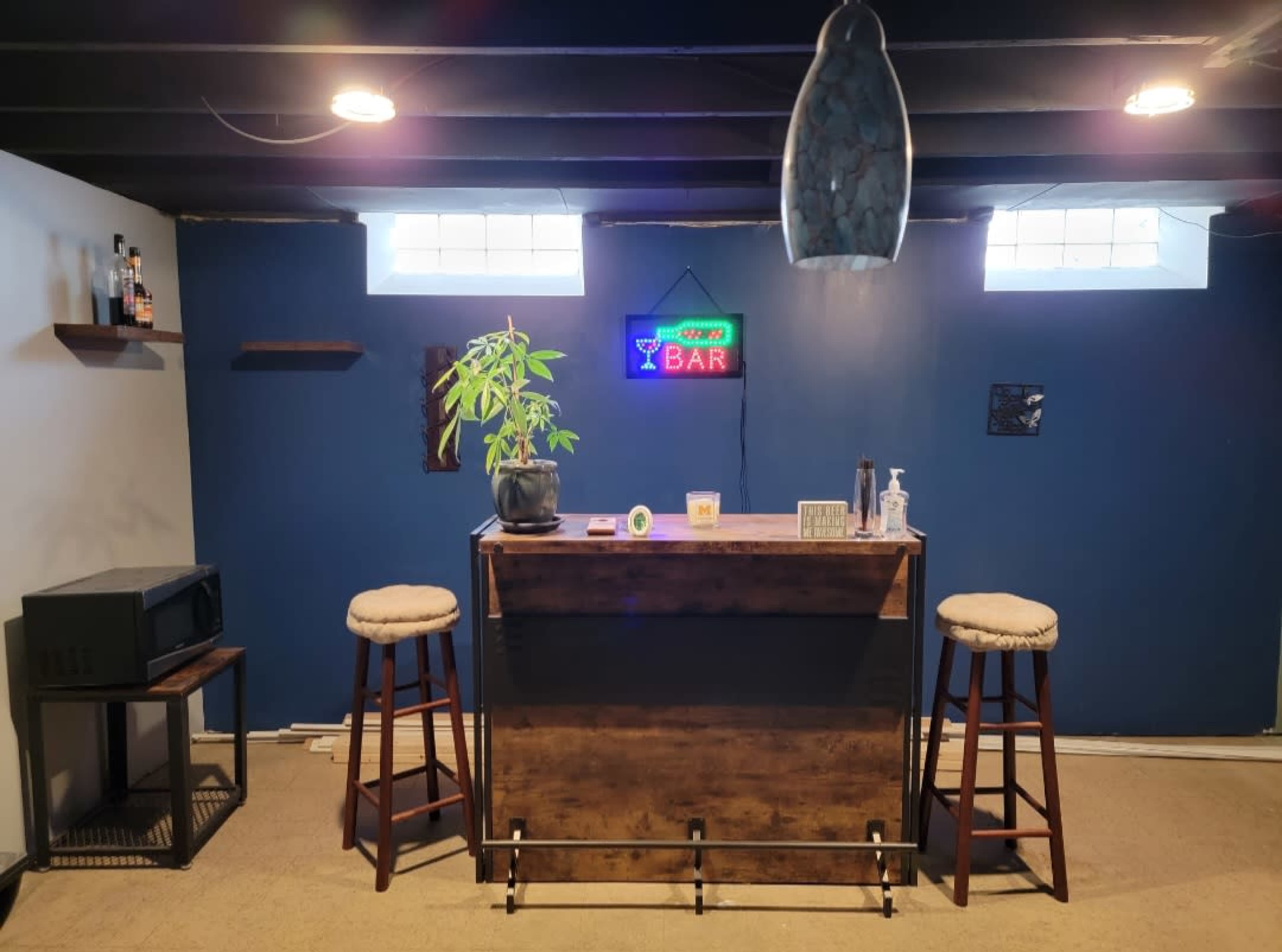 The image shows a small home bar area with a wooden bar counter, two stools, a microwave, and a neon "BAR" sign, all set against a blue wall.