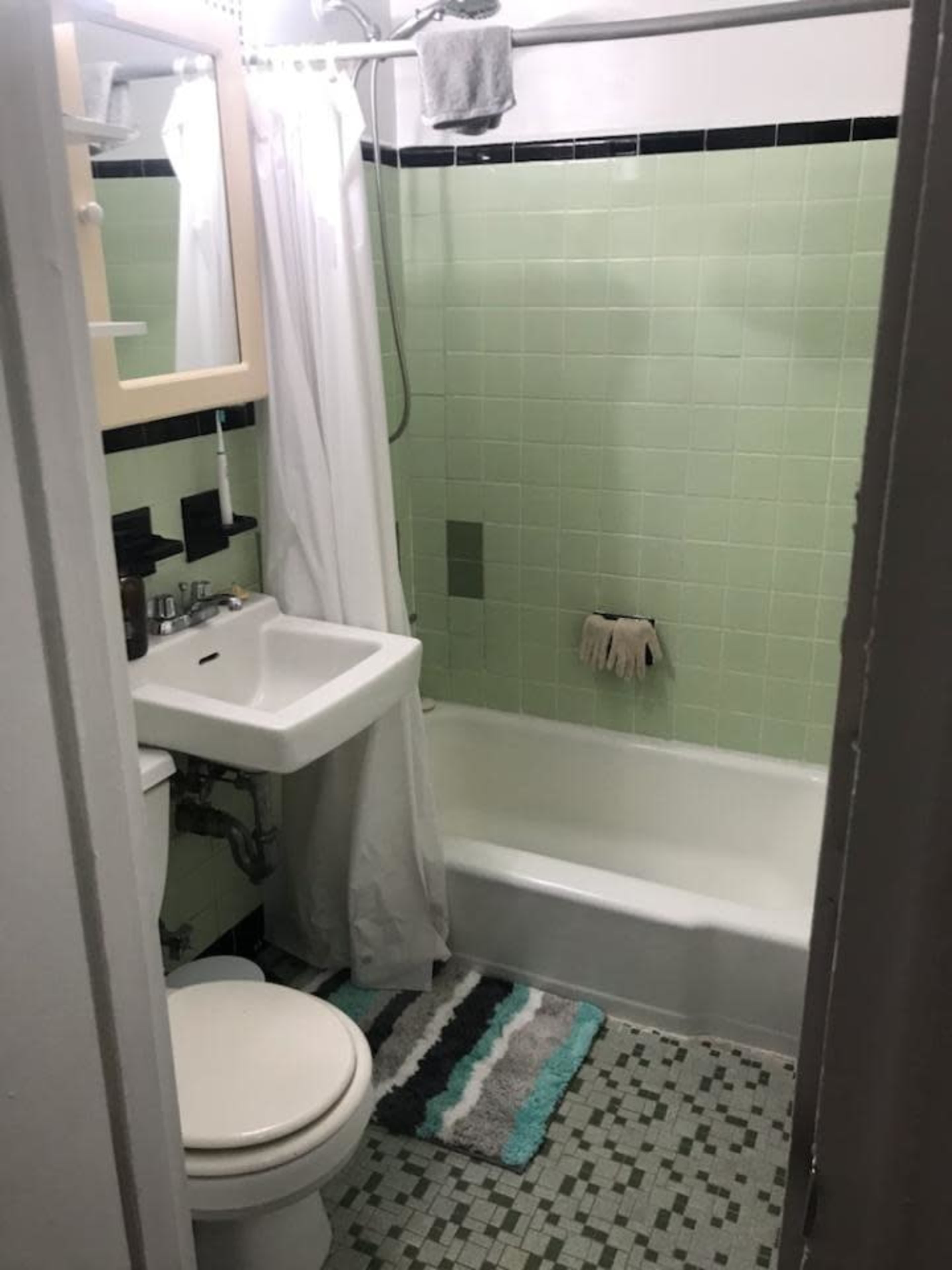 A small bathroom features a bathtub, toilet, and sink with green tile walls and a shower curtain.