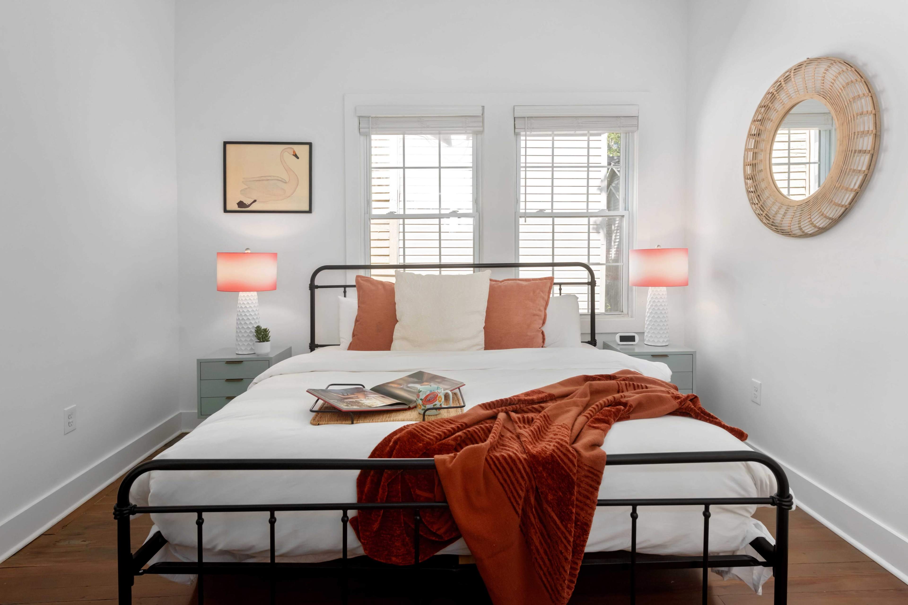The image shows a neatly organized bedroom with a metal bed frame, white linens, and decorative pillows, two bedside lamps, and artwork on the walls.