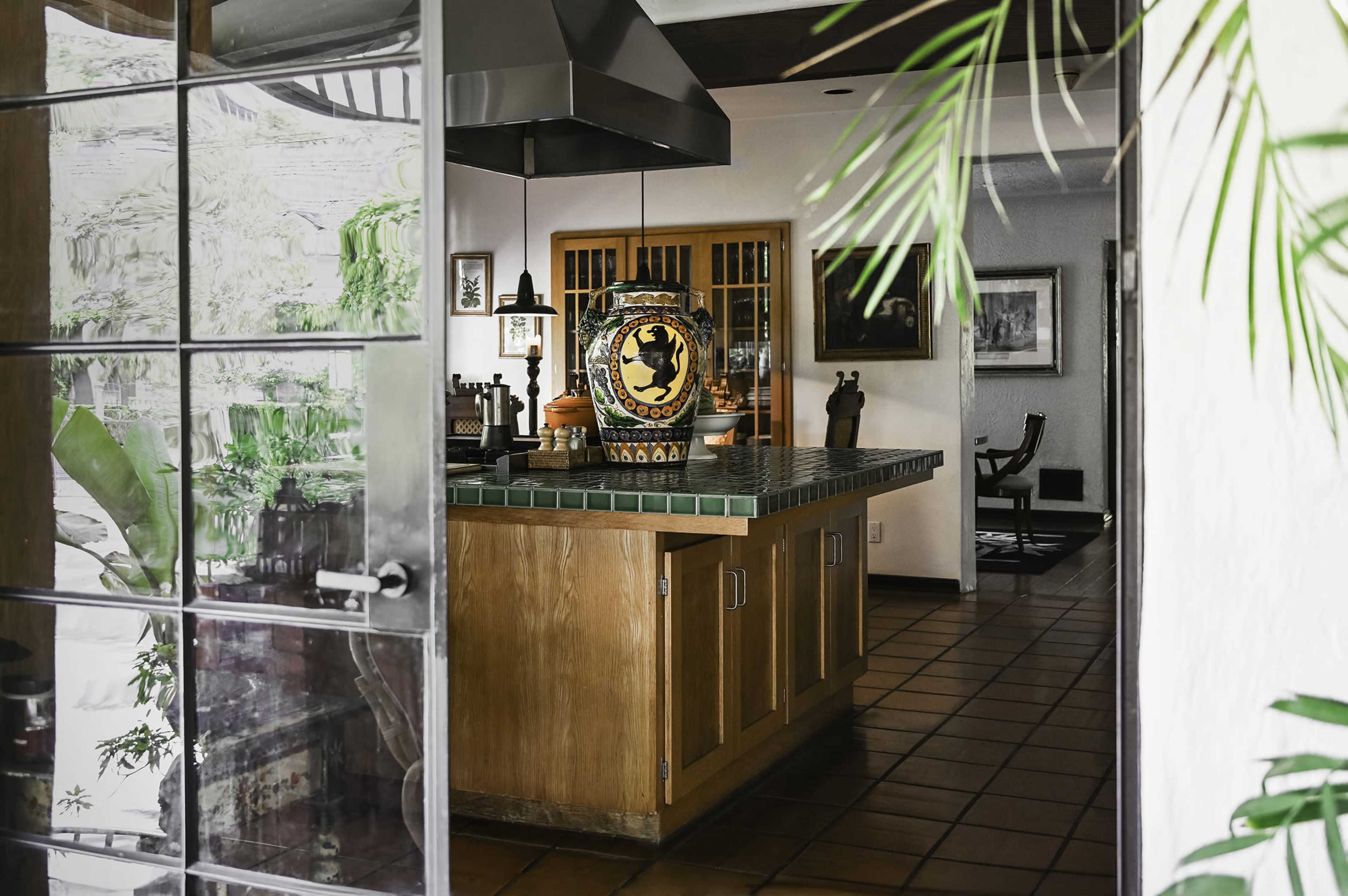 A spacious kitchen with wooden cabinets, a tiled island, and a decorative vase, visible through a glass door framed by greenery.