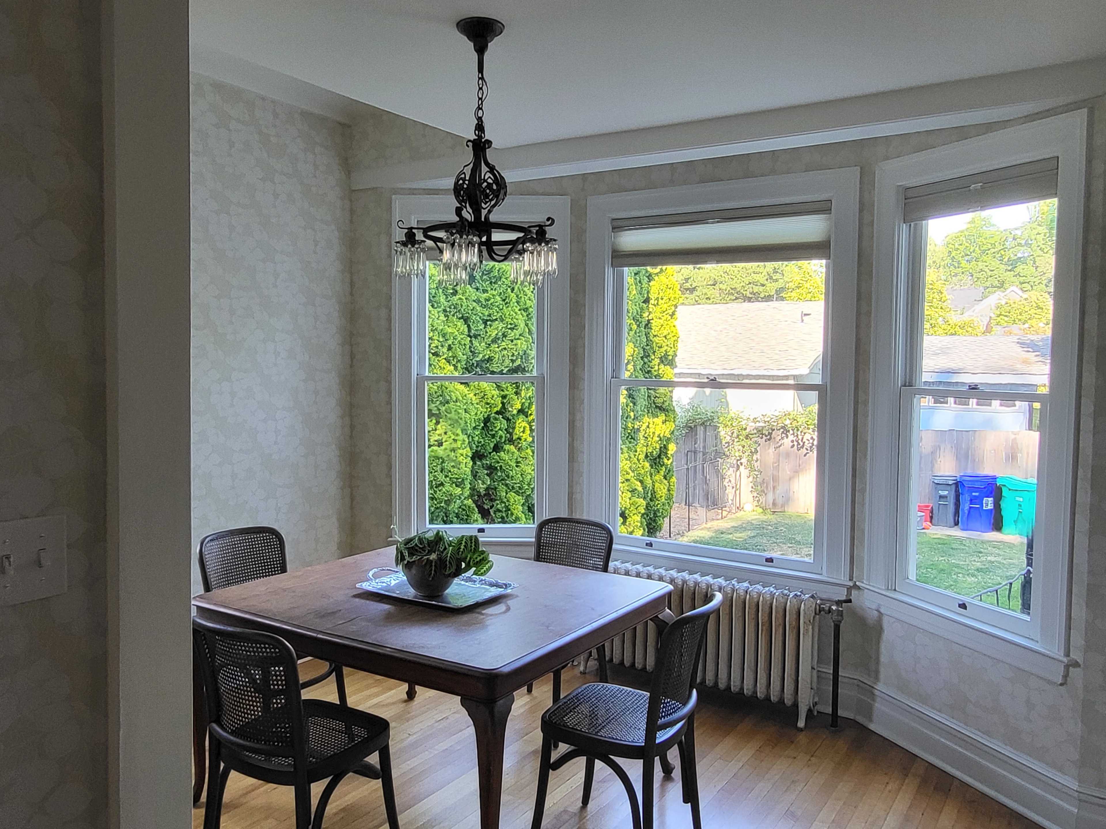 A dining area features a wooden table surrounded by four chairs, with large windows providing a view of greenery outside.