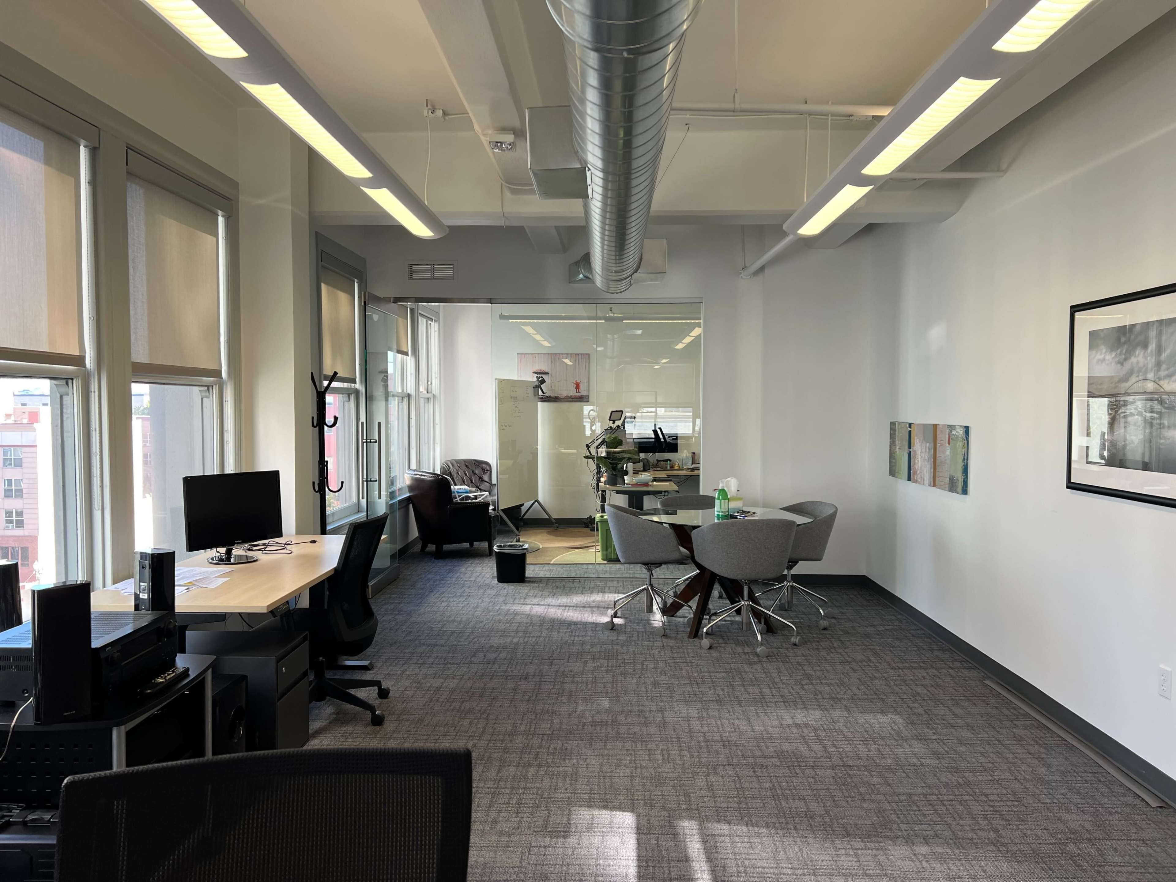 A modern office space featuring desks, chairs, and a conference area with large windows and light-colored walls.