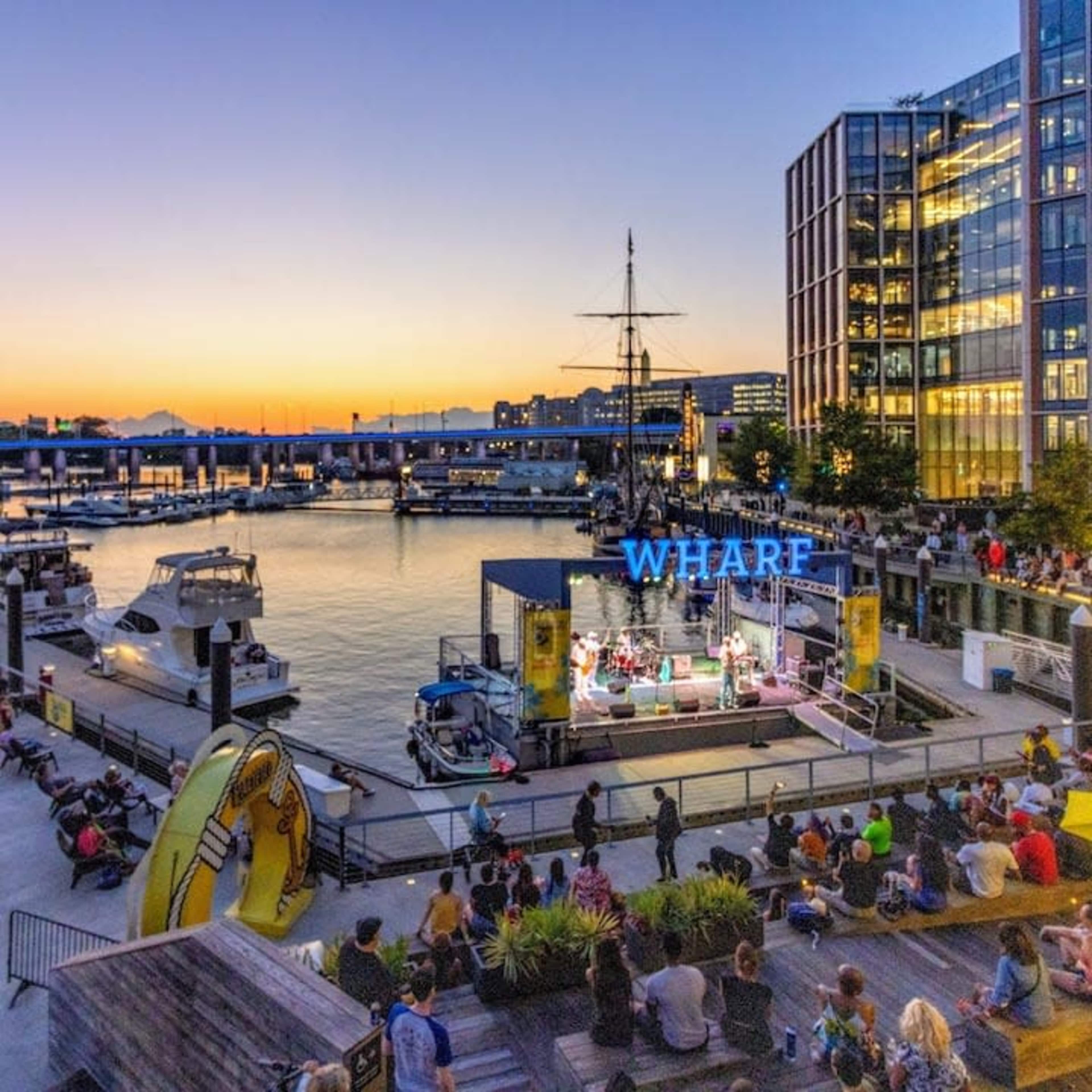 A crowd gathers at the Wharf to enjoy a live music performance as boats drift in the harbor during sunset.