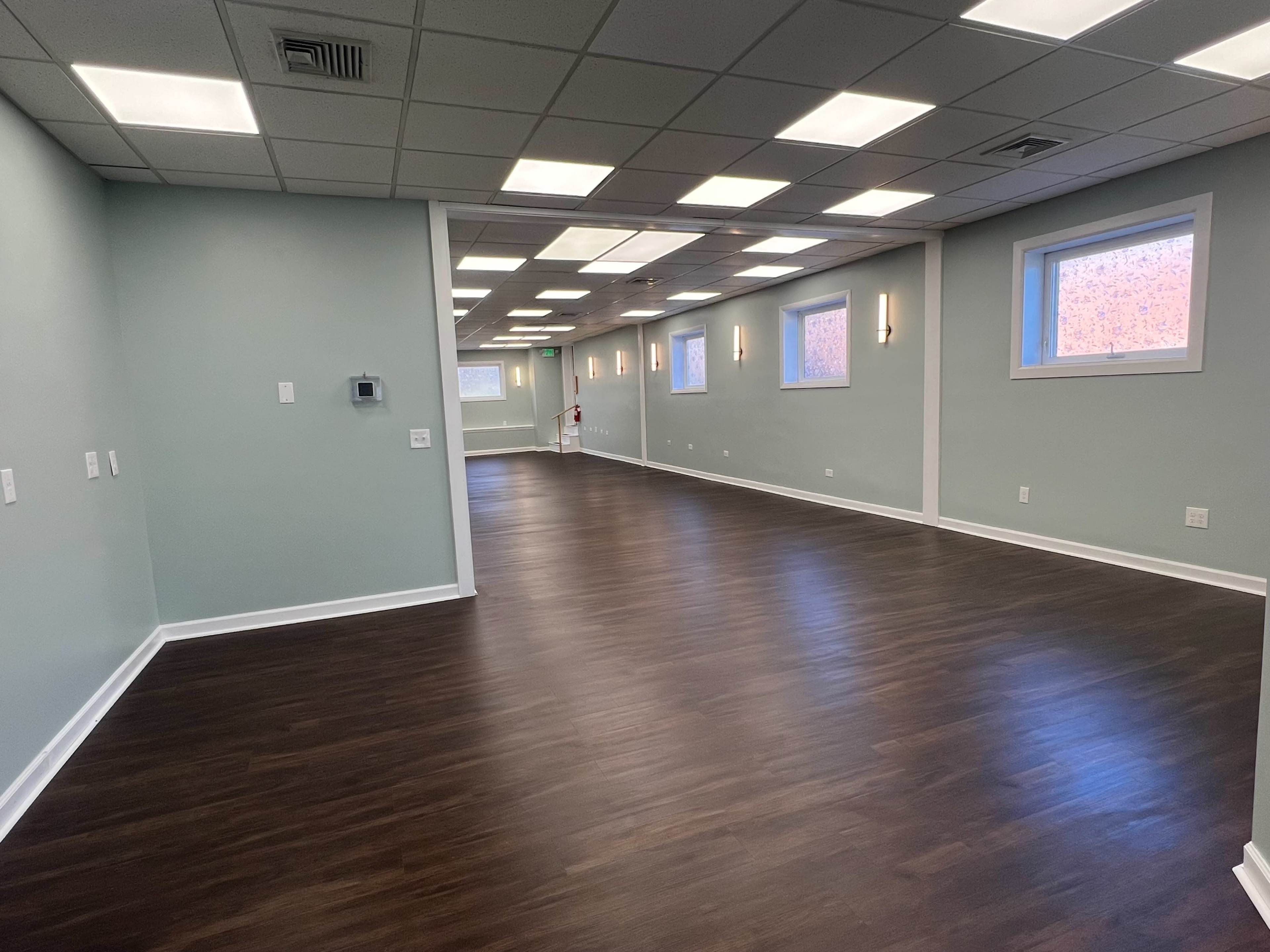 The image shows an empty, well-lit room with wooden flooring and light green walls, featuring several windows and wall sconces.