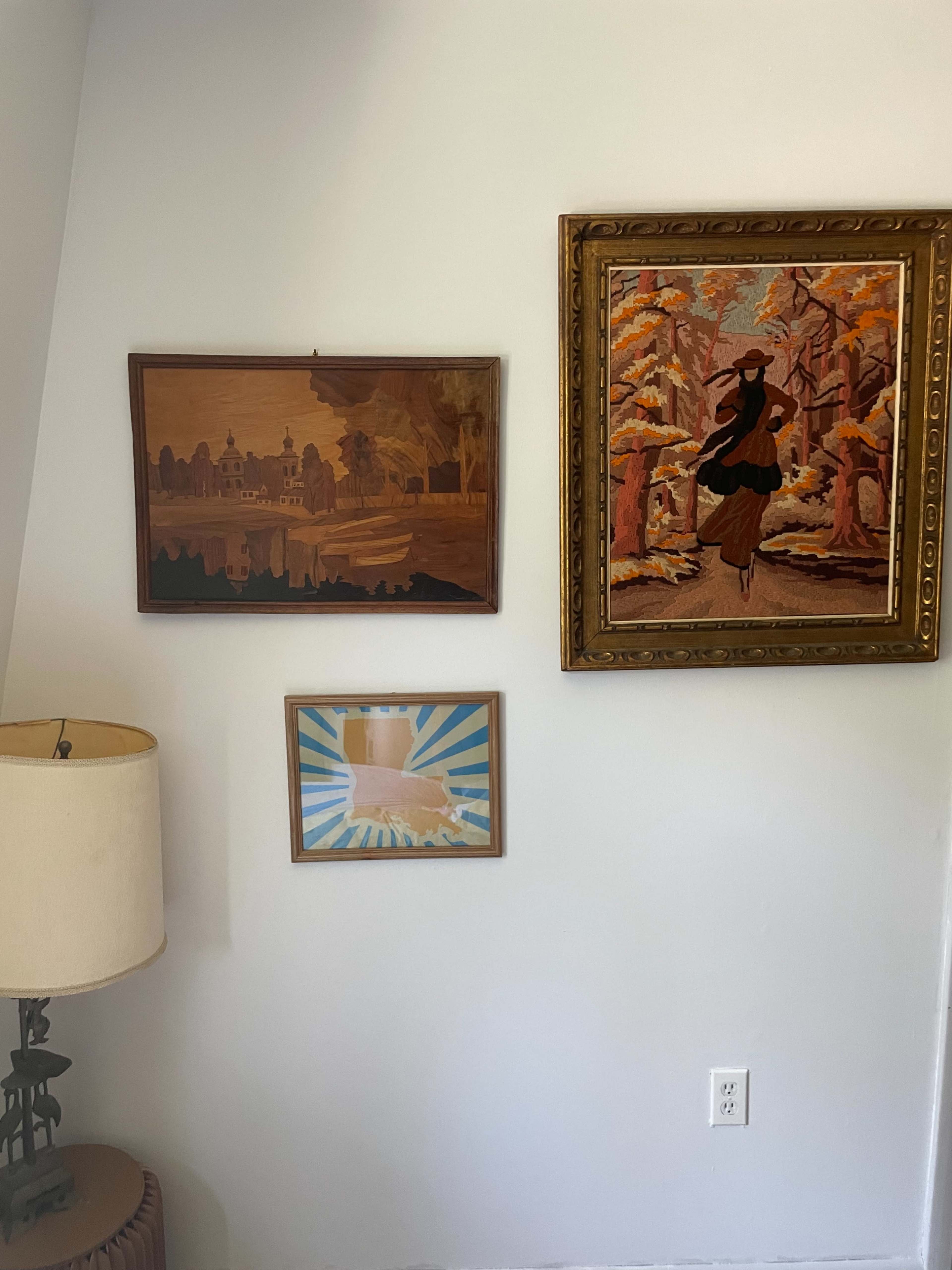 A corner of a room with a lamp, three framed artworks on the wall, and a white outlet.