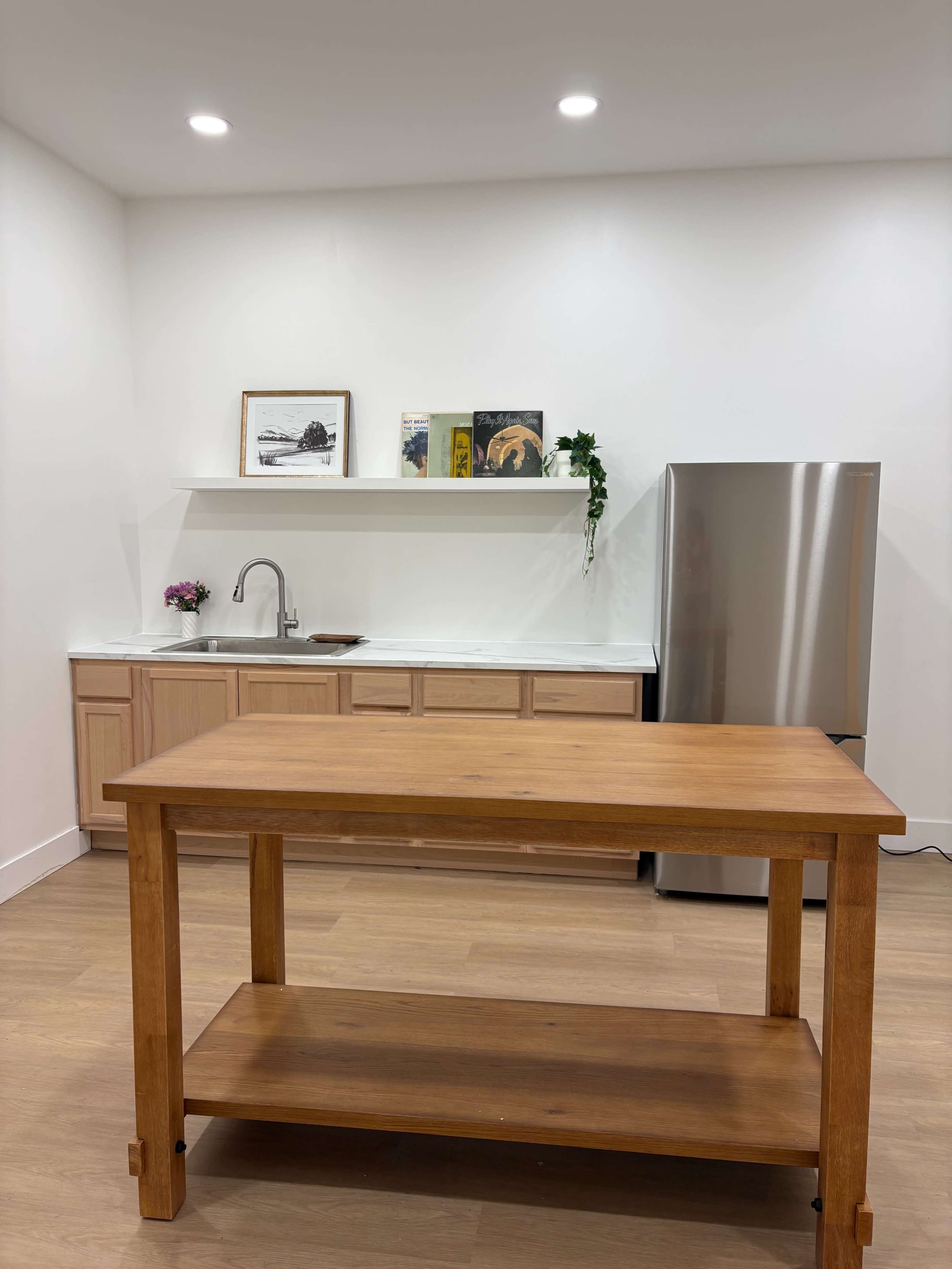 The image shows a modern kitchen featuring a wooden island table, a stainless steel refrigerator, a sink with a faucet, and a shelf with decorative items on the wall.