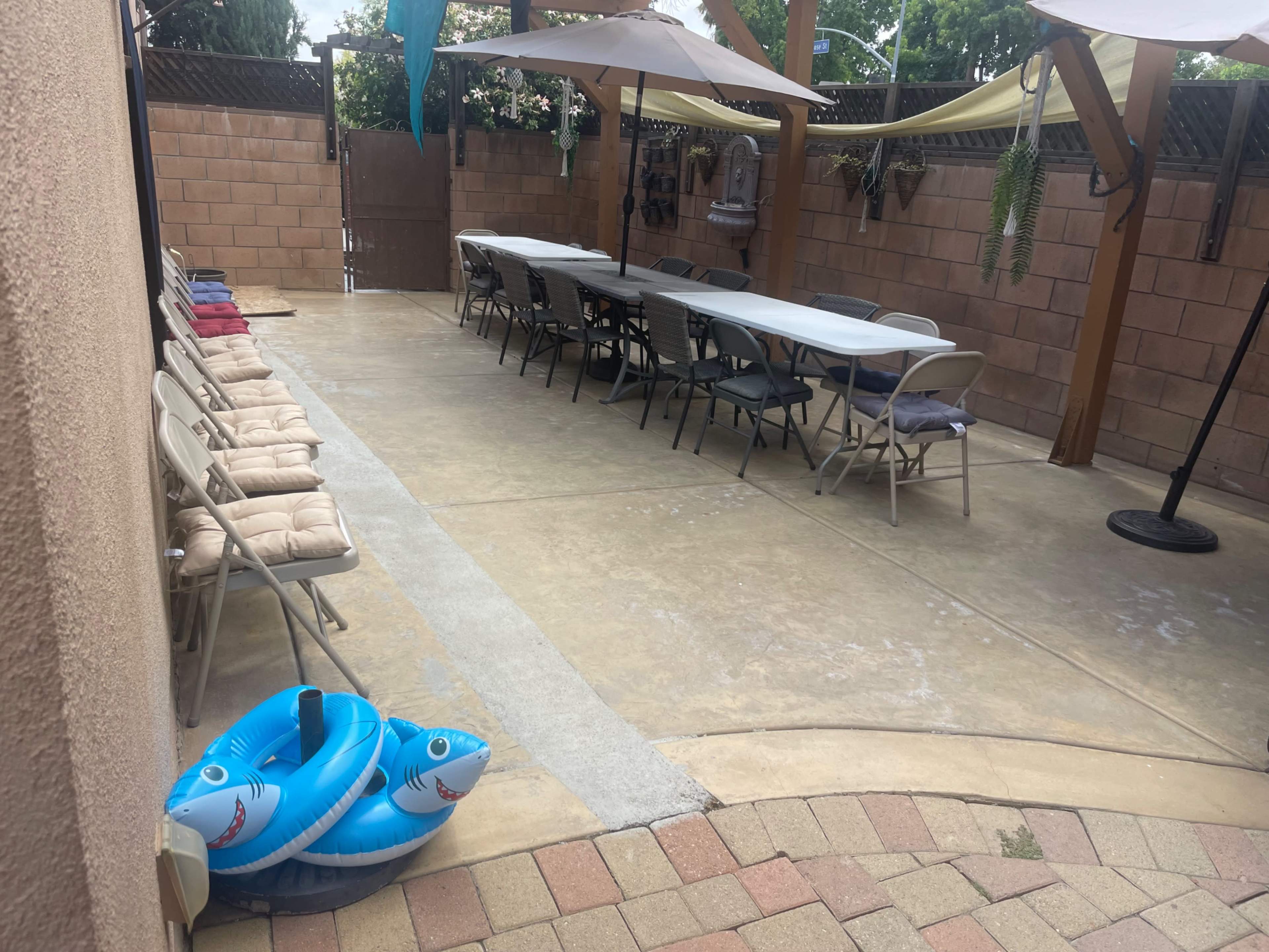 The image shows a patio area with a long table set for seating, surrounded by folding chairs, and two blue inflatable pool toys resting on the ground.
