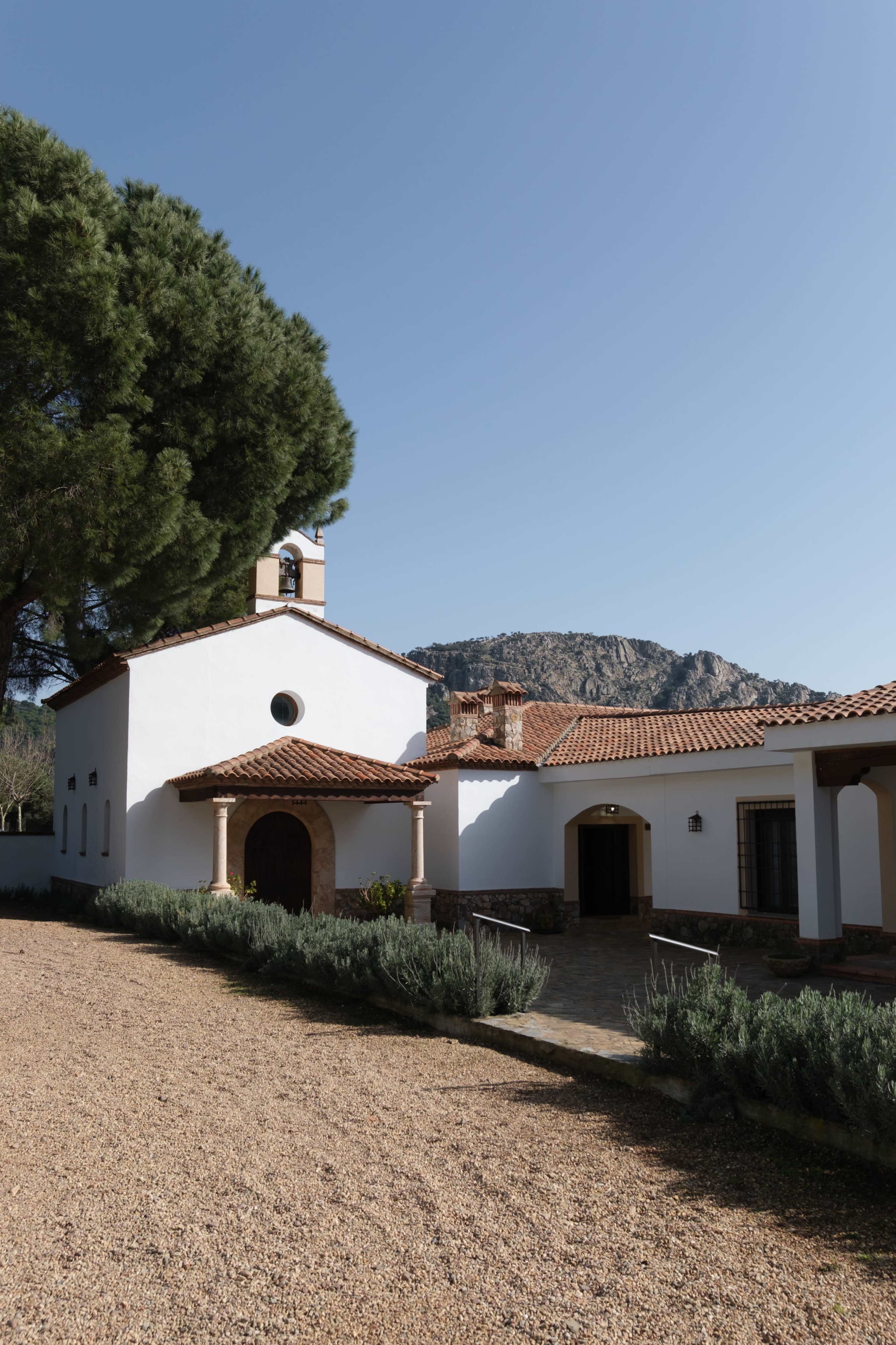 A white stucco house with a red-tiled roof and a bell tower is situated near a mountain, surrounded by gravel landscaping and greenery.
