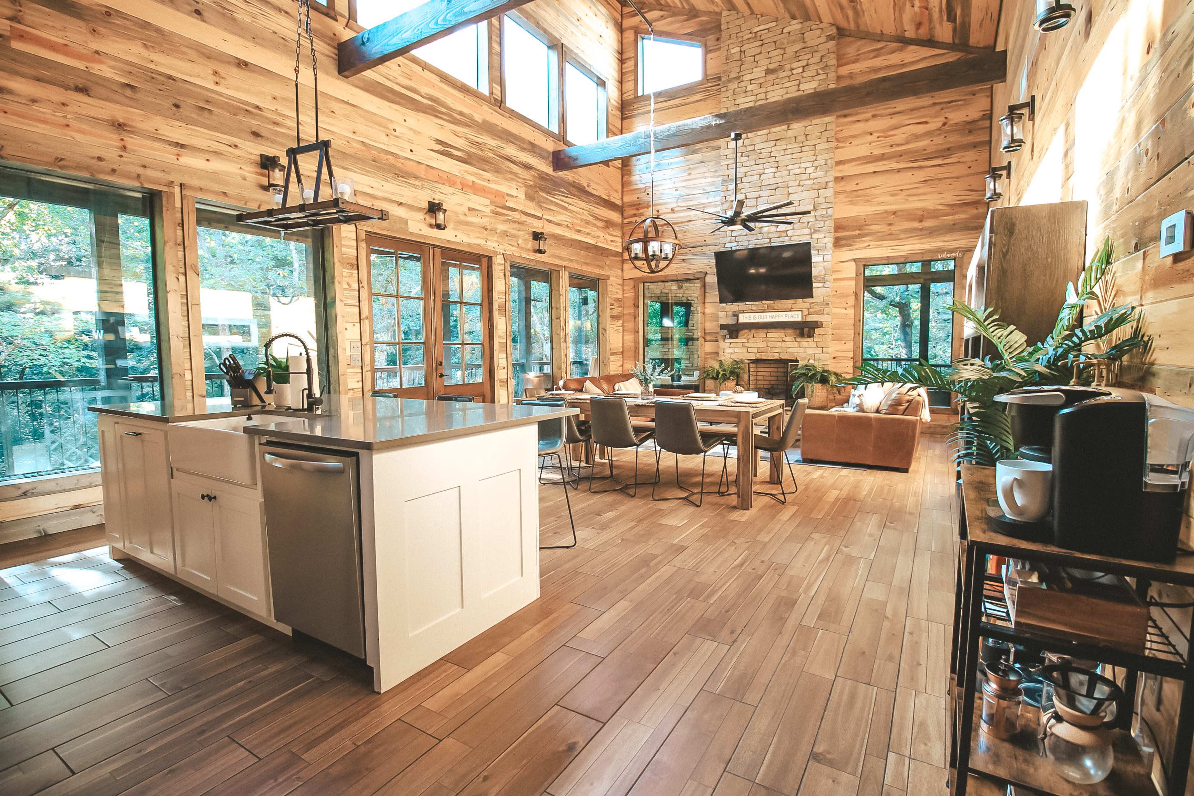 A spacious, modern cabin interior featuring a combined kitchen and living area with high wooden ceilings, large windows, and a stone fireplace.