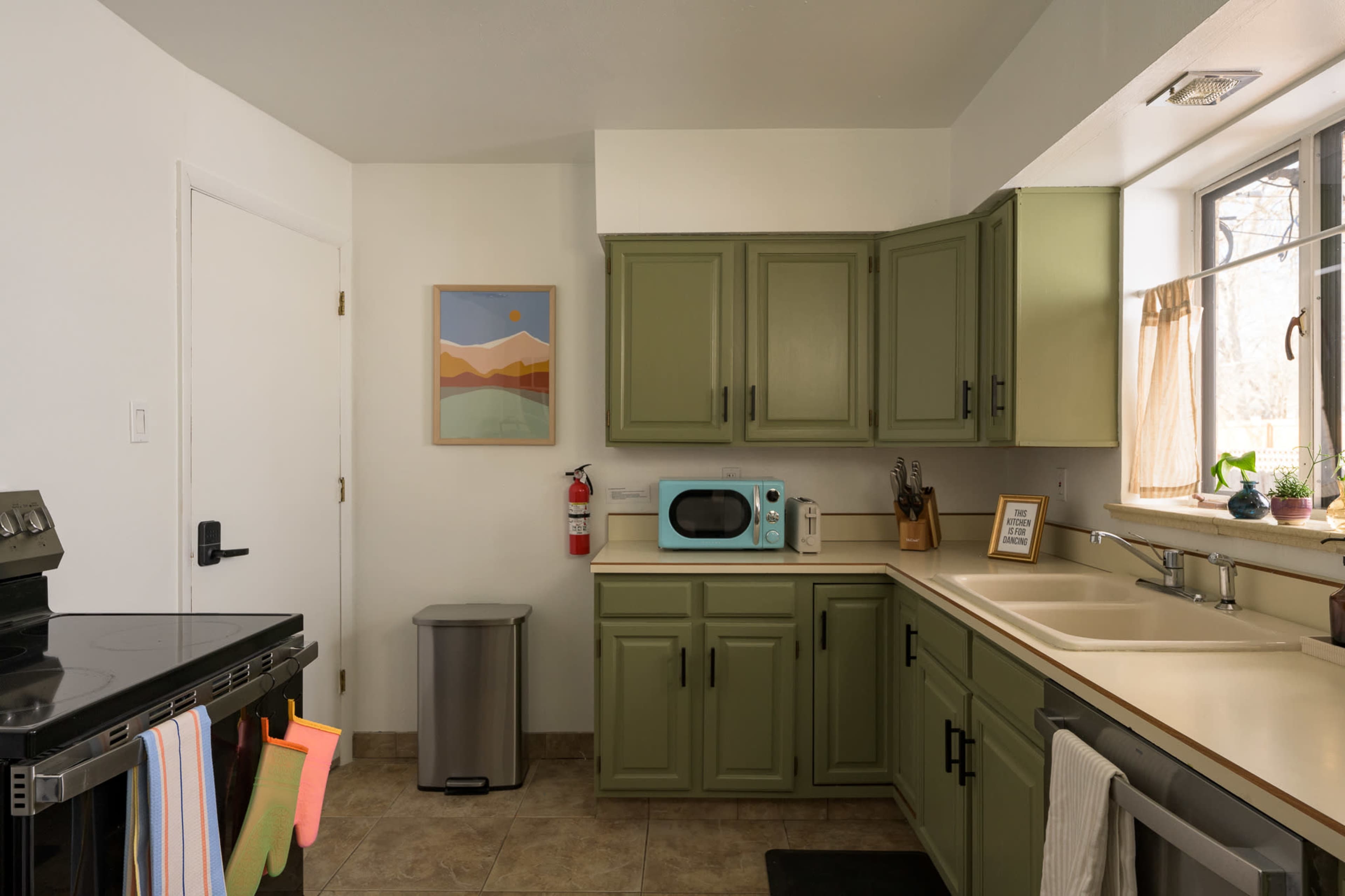 The kitchen features green cabinets, a black stove, a white sink, and a turquoise microwave, with a window and a door visible.