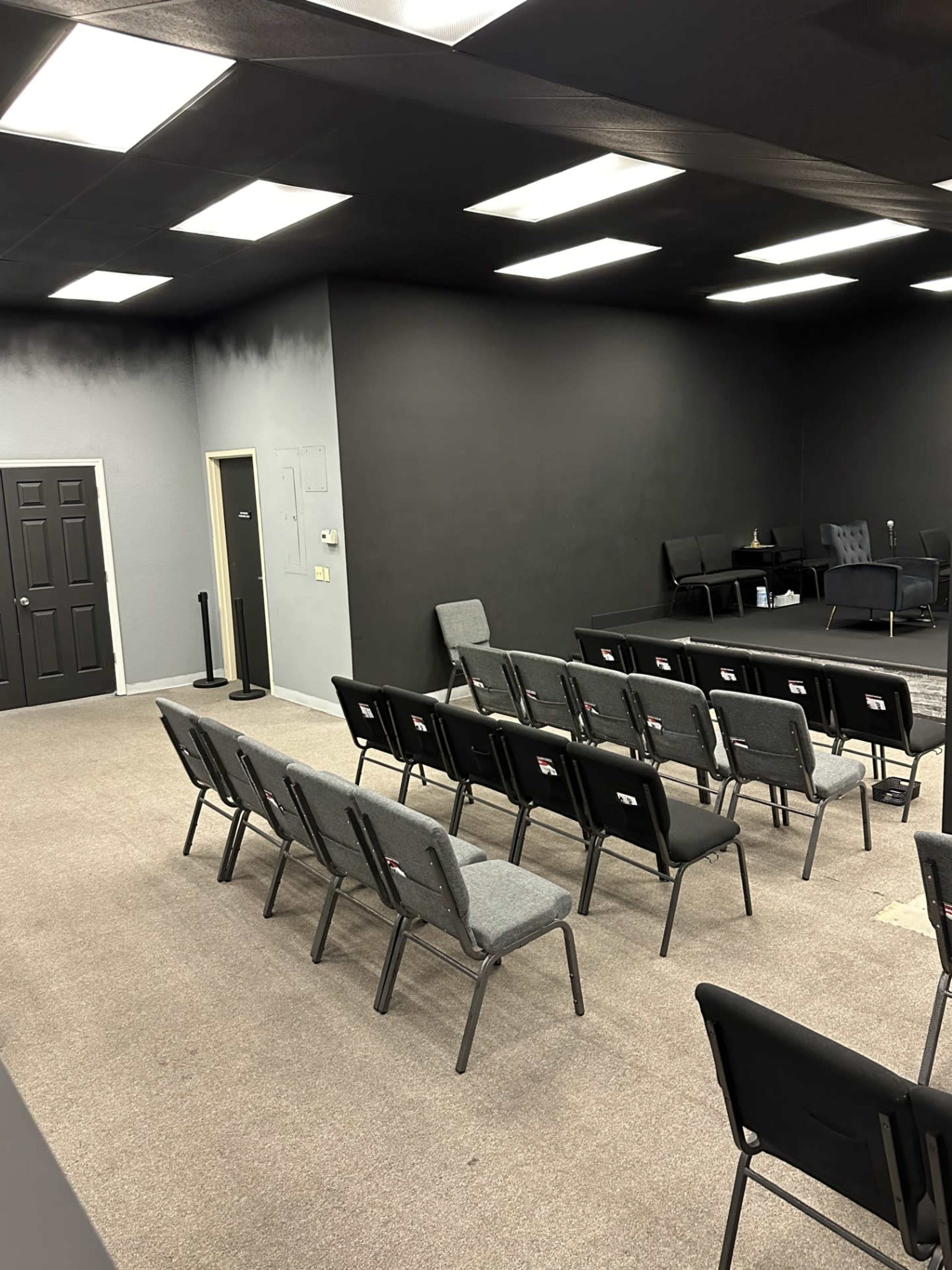 A sparsely furnished room with rows of gray chairs facing a black wall, and a small seating area in the background.