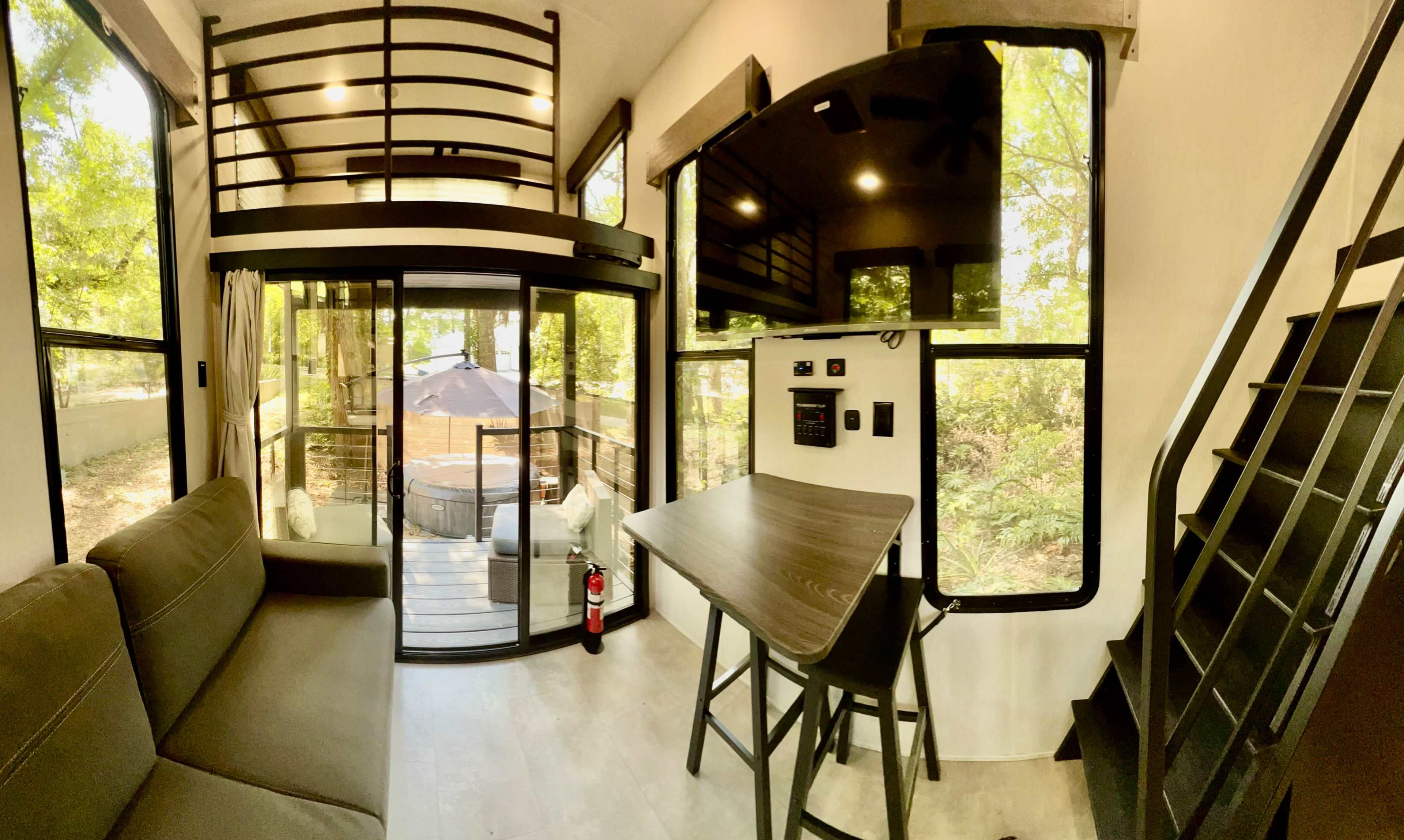 The interior of a modern tiny house features a living area with a sofa, a table with stools, and large windows overlooking a deck surrounded by trees.