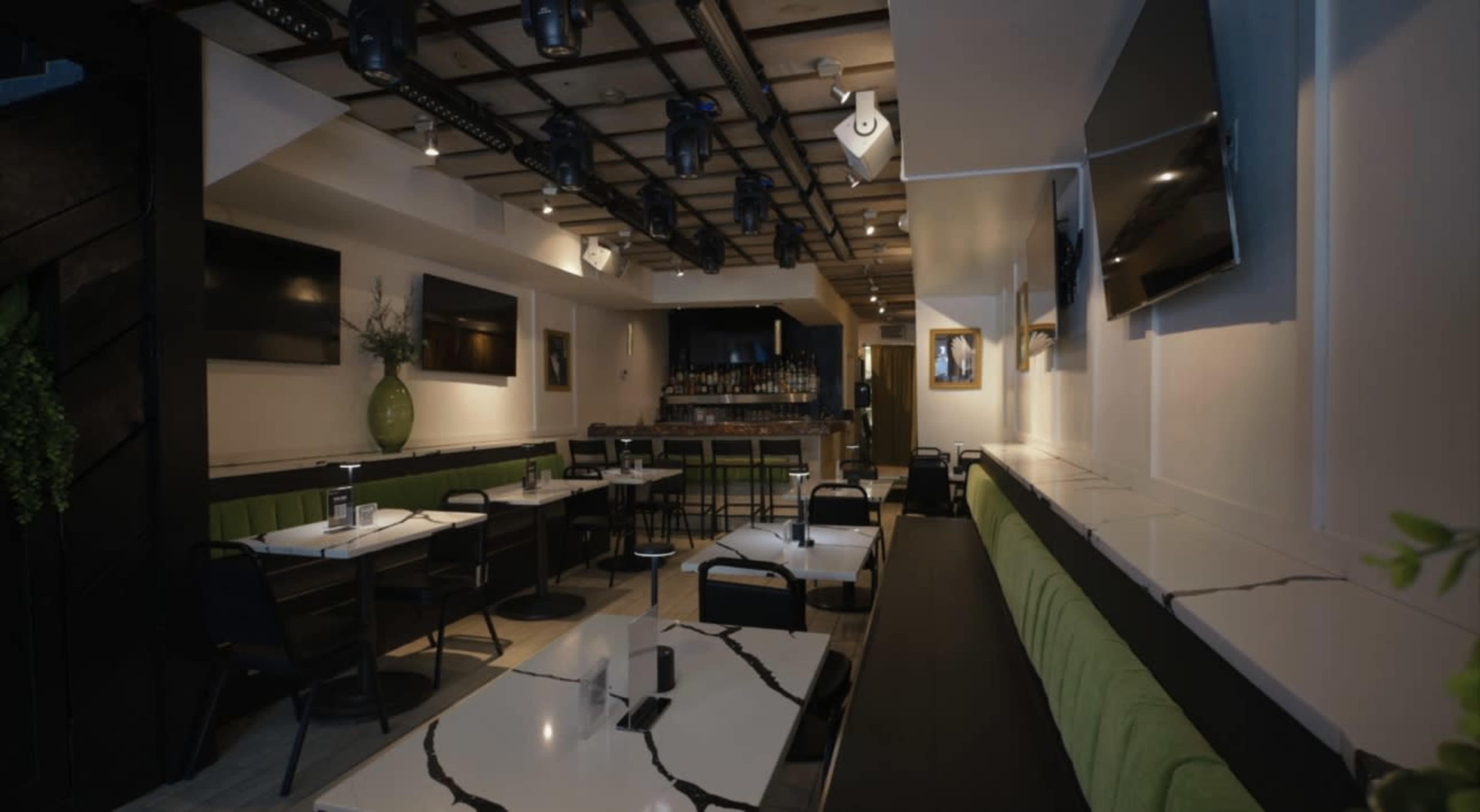 The image shows an interior view of a modern restaurant featuring green-upholstered seating, marble-patterned tables, and a well-lit bar area in the background.