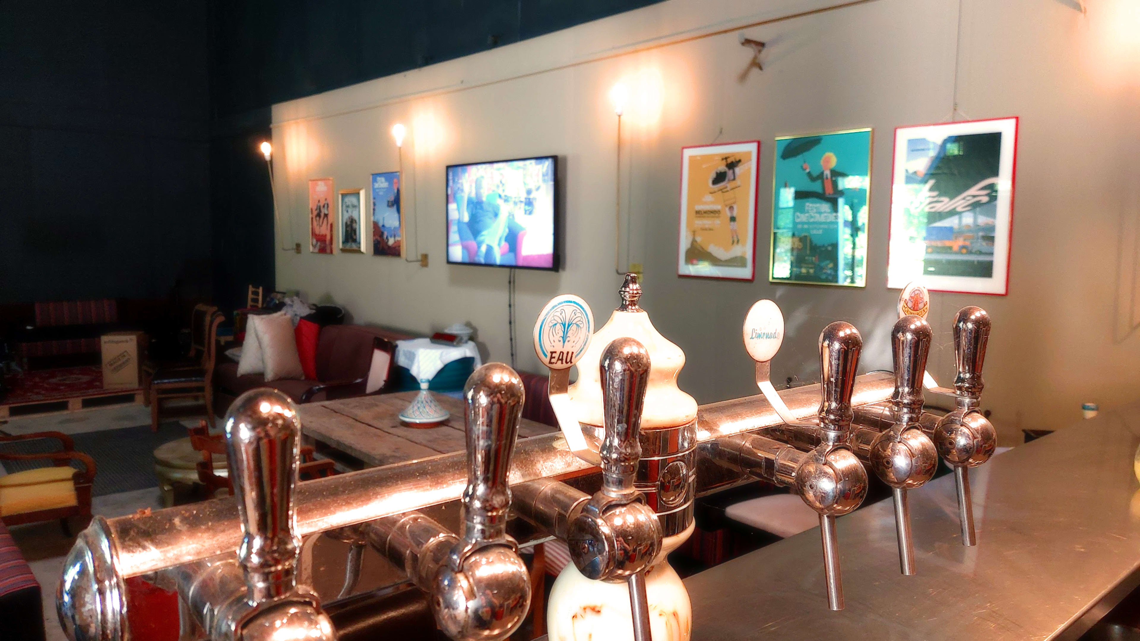 The image shows a bar area with a row of beer taps in the foreground and a lounge-like seating arrangement with posters and a television on the wall in the background.