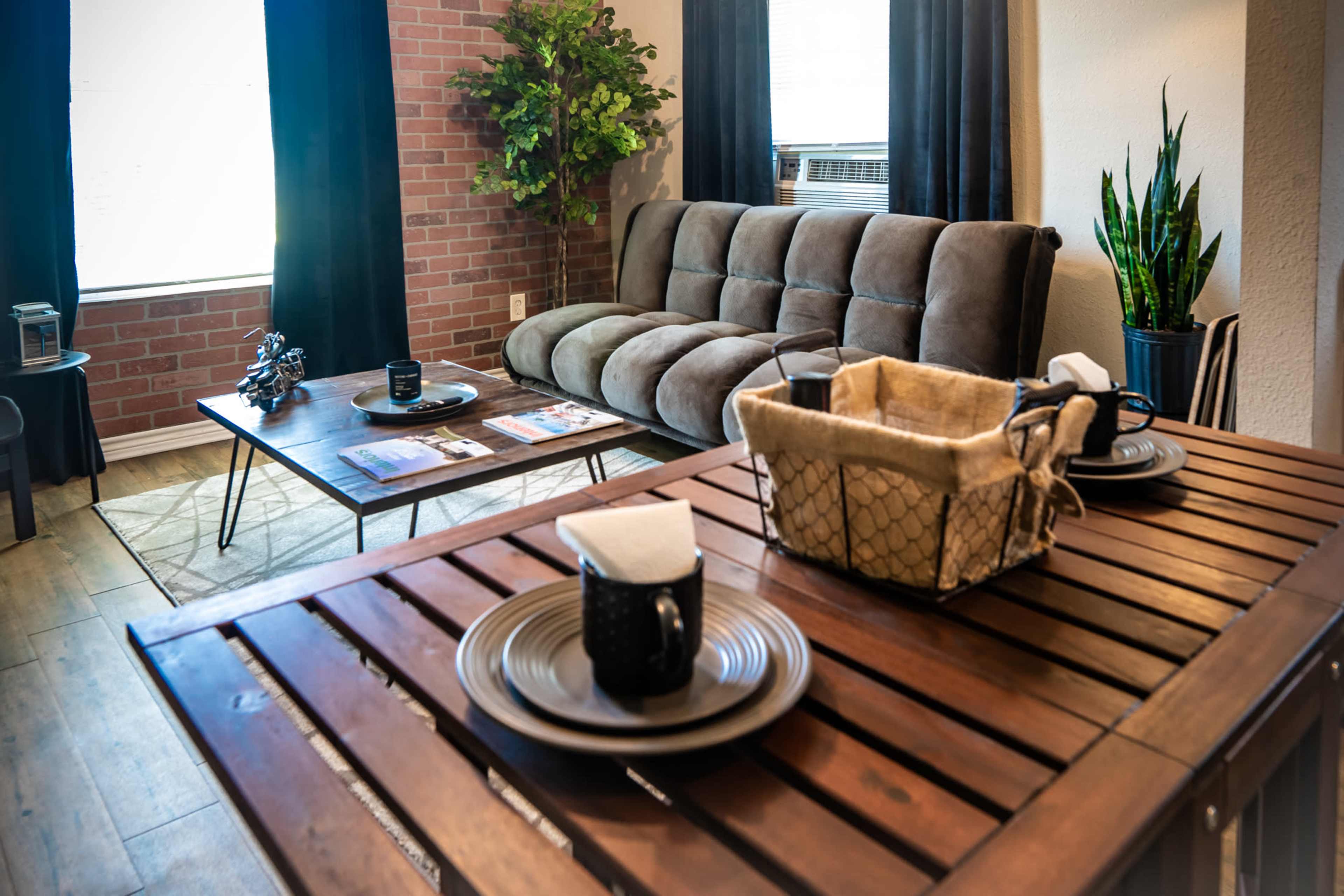 A cozy living room with a brown sofa, a wooden table set for dining, and decorative plants in the corners.