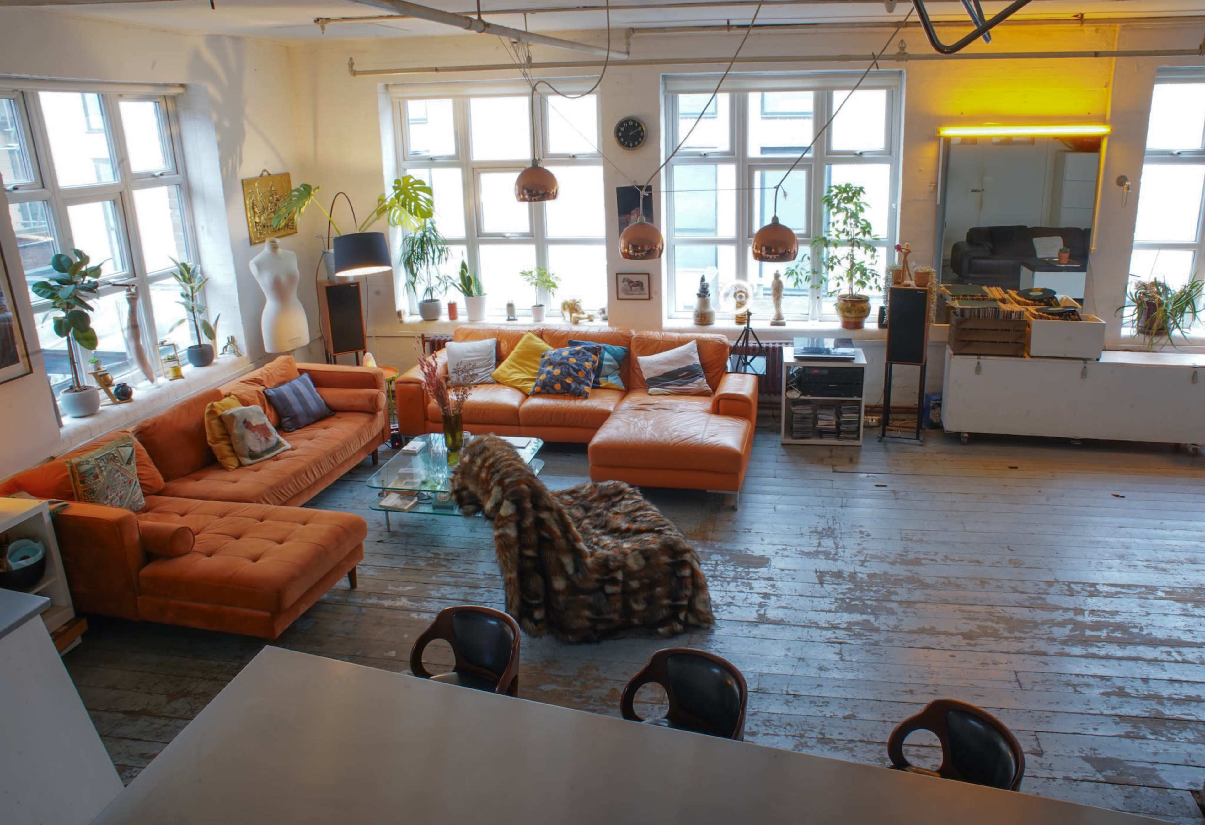 A spacious living area features a large orange sectional sofa, a glass coffee table, and several indoor plants, illuminated by natural light from multiple windows.