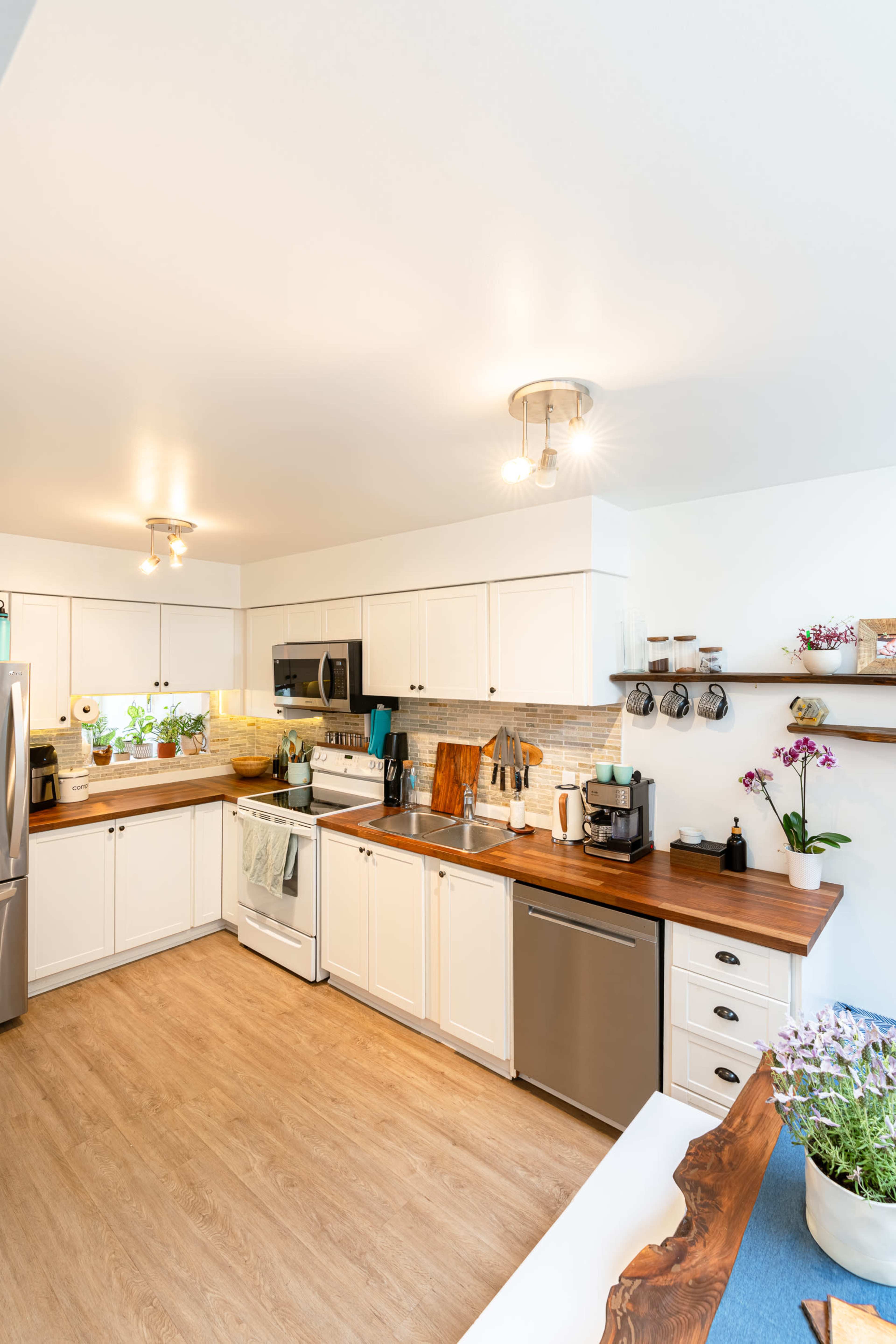 A modern kitchen features white cabinetry, stainless steel appliances, wooden countertops, and potted plants on the windowsill.
