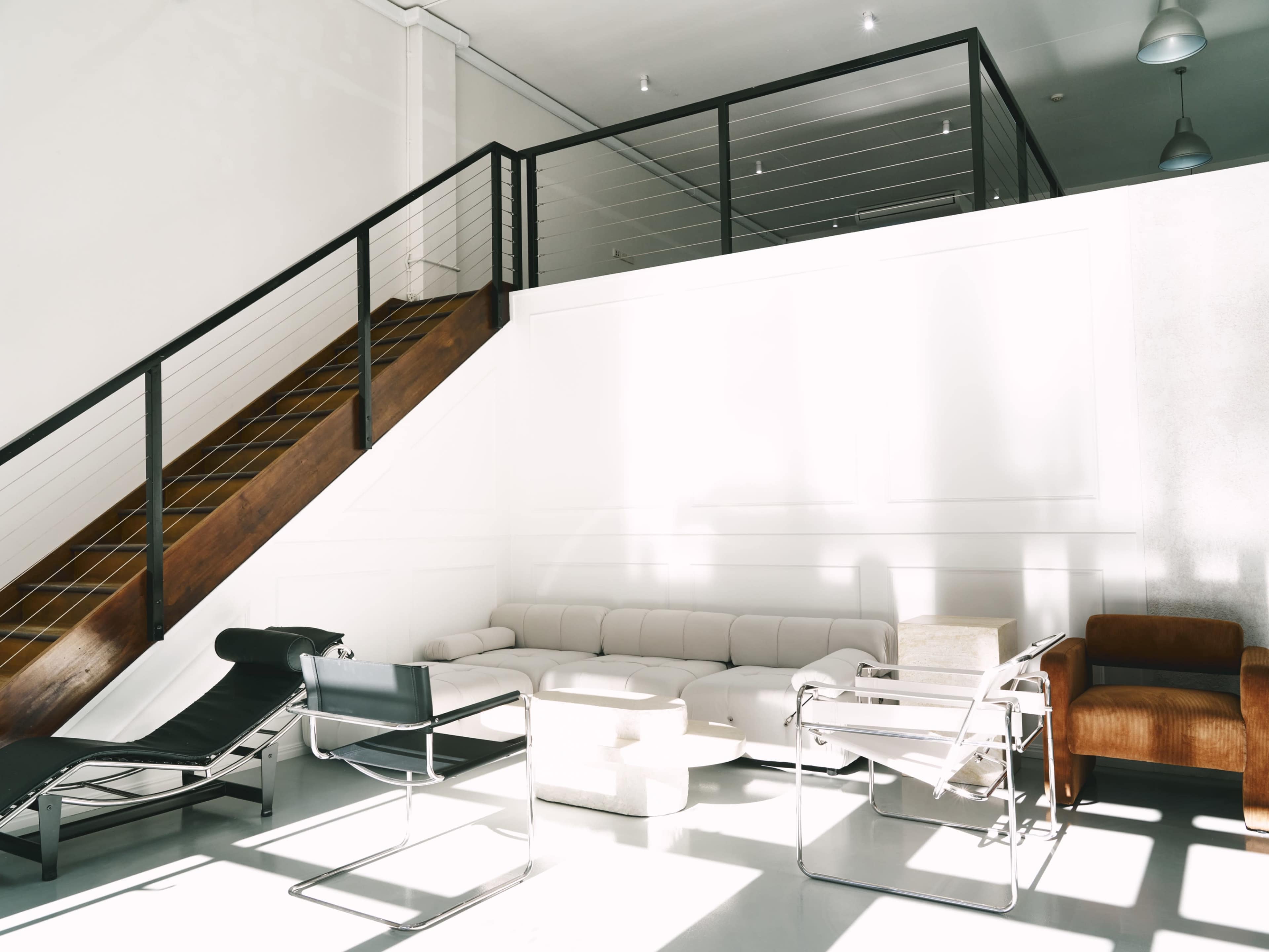 The image shows a modern, minimalist interior space with a staircase, a sectional sofa, and various lounge chairs.